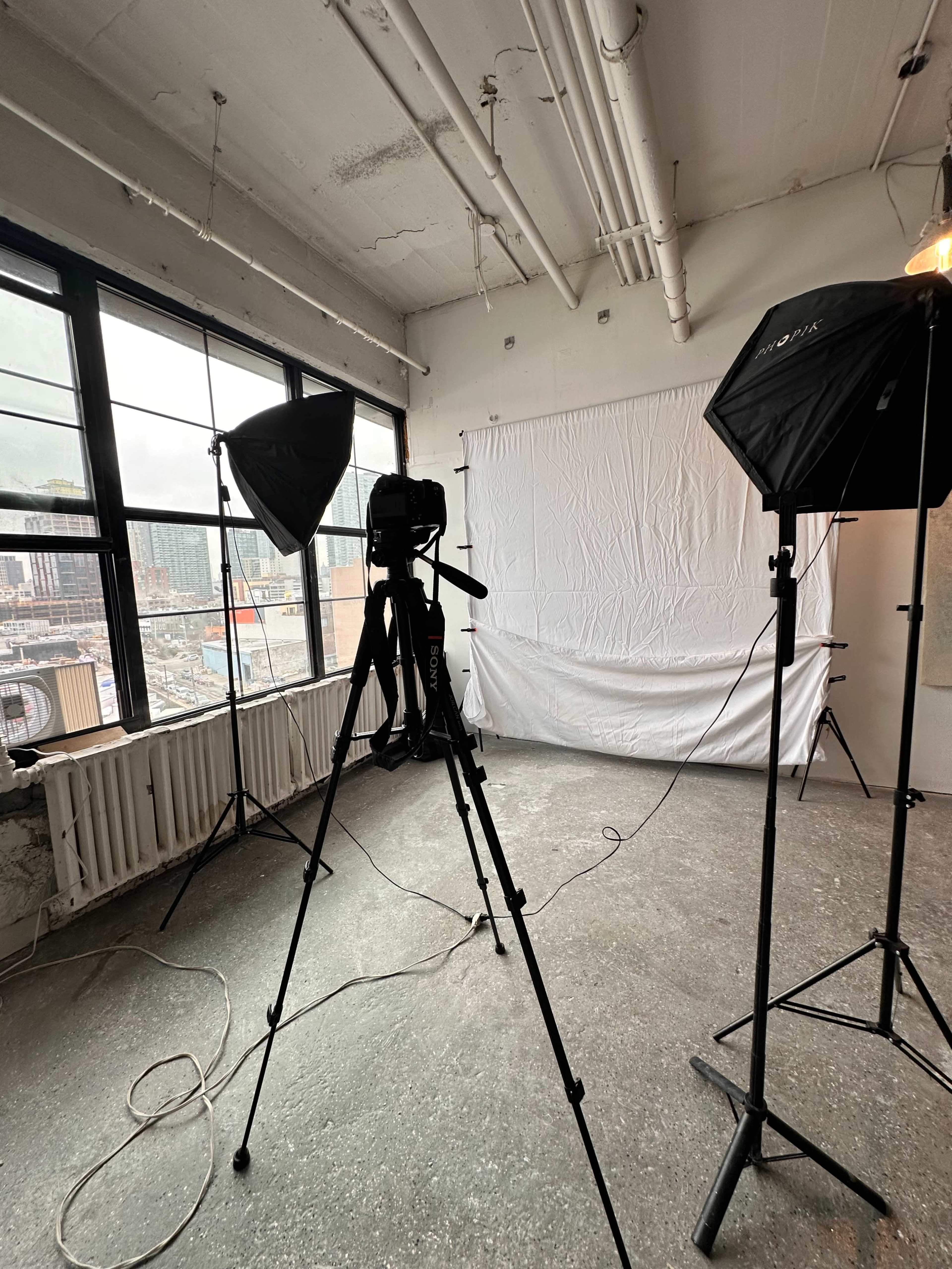 The image shows a photography setup in a room featuring a camera on a tripod, two softbox lights, and a white backdrop facing large windows.