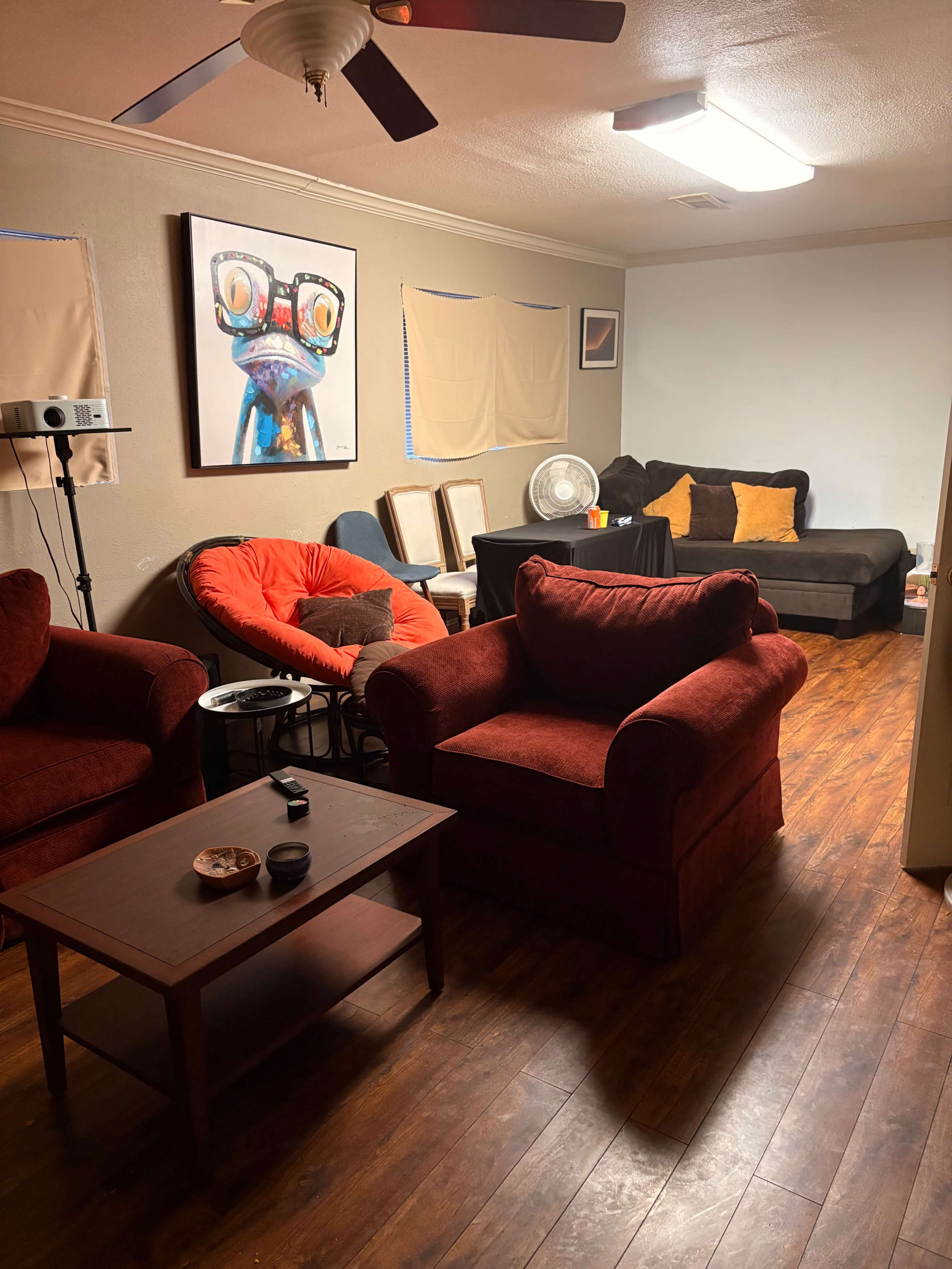 The image shows a living room with a red sofa and chairs, a dark couch, a coffee table, and a colorful piece of wall art.