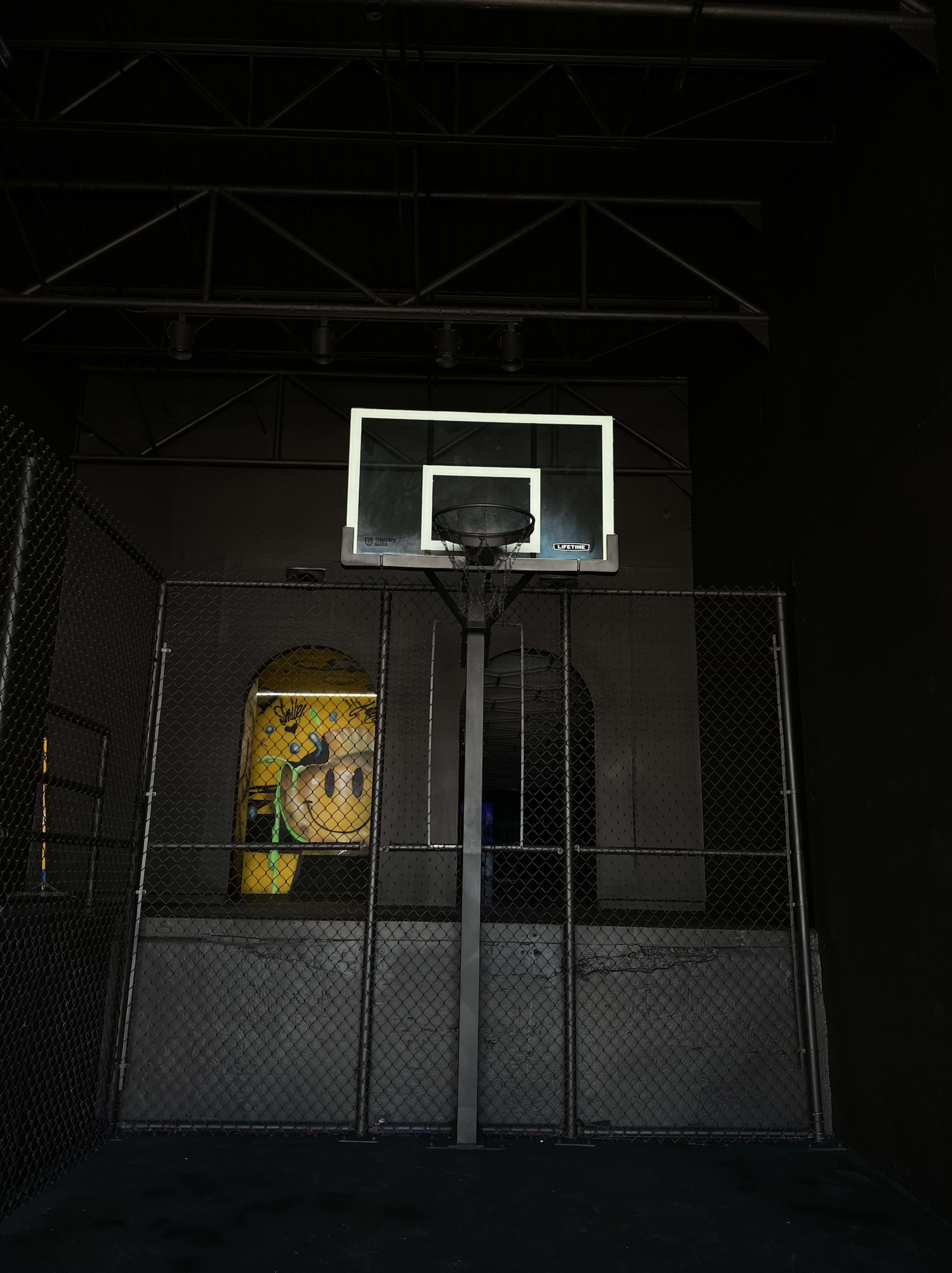 A basketball hoop installed in a dimly lit indoor court enclosed by a chain-link fence, with a large mural partially visible in the background.