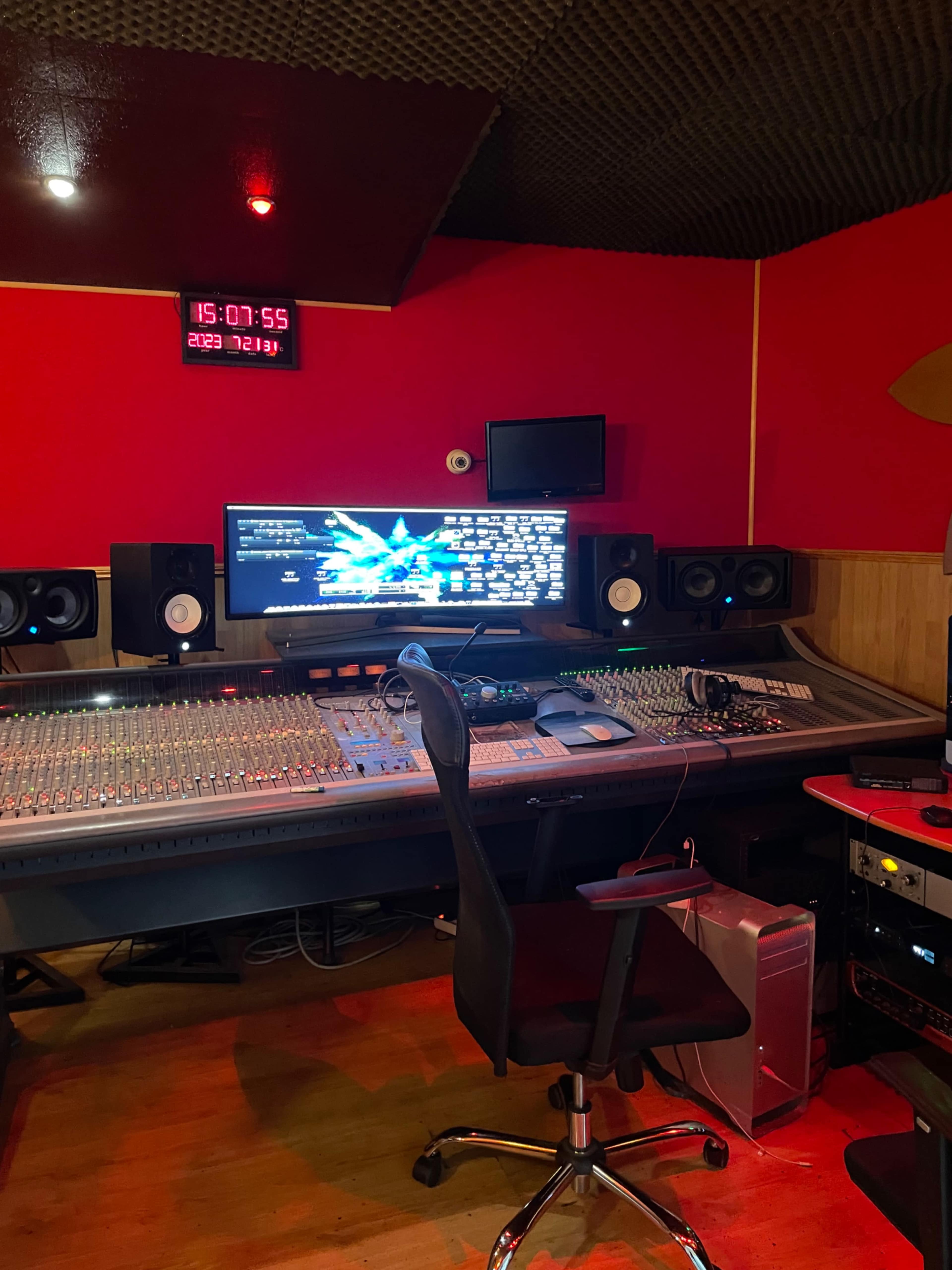 A music studio featuring a large mixing console, computer monitor, and sound equipment against a red wall and acoustic paneling.