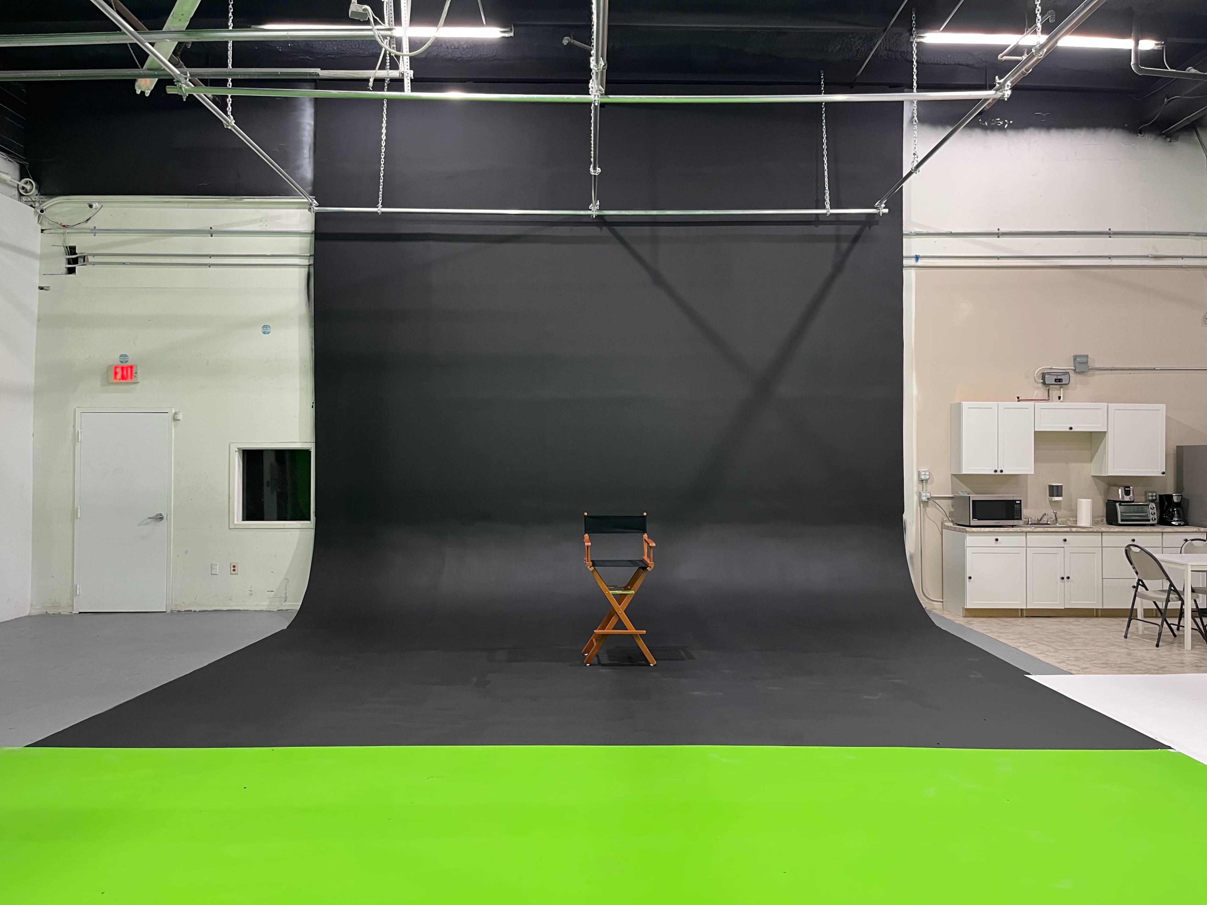 The image shows a photography studio featuring a large black backdrop, a green floor section, and a wooden director's chair positioned in front of the backdrop.