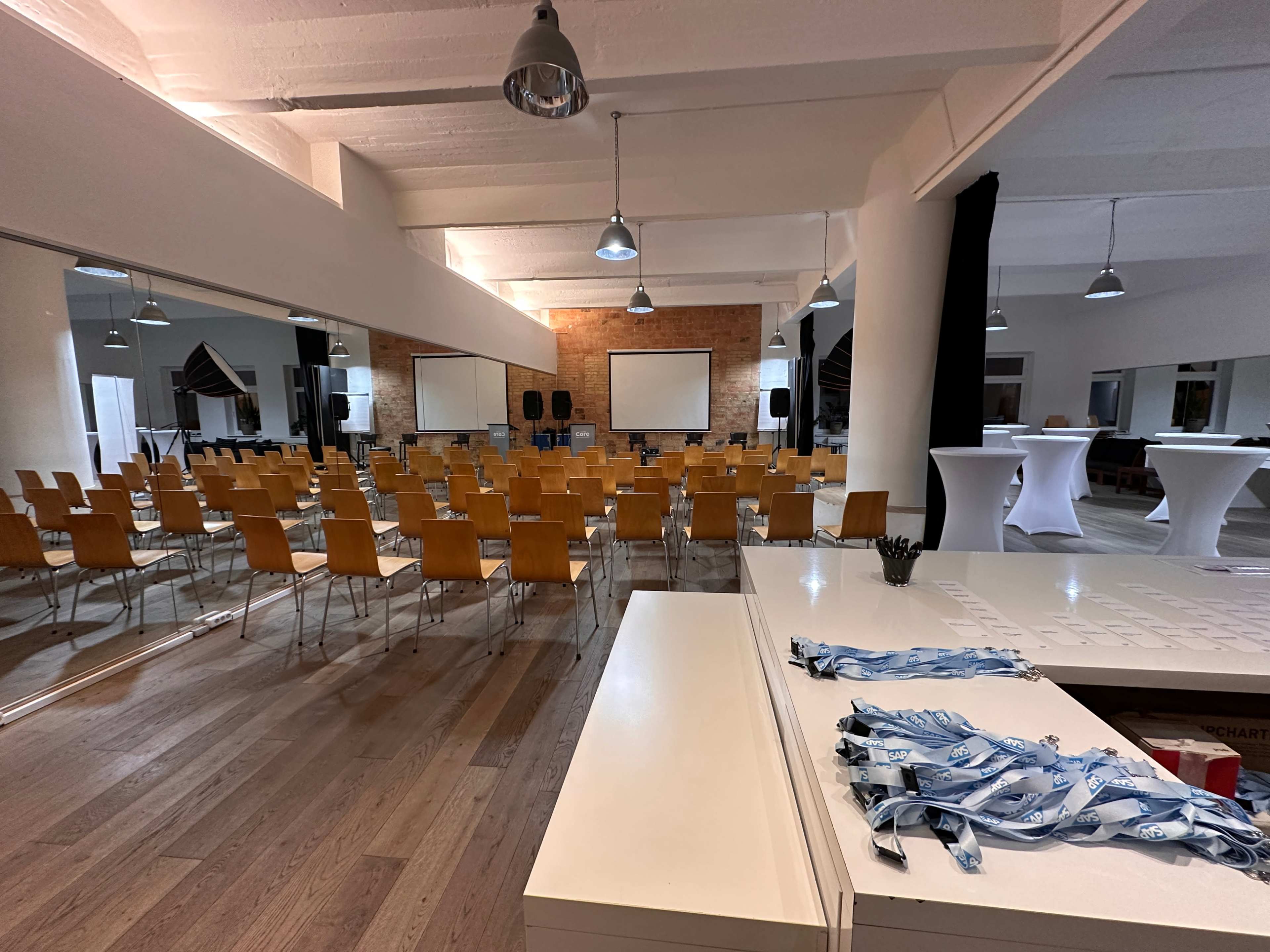 The image shows a spacious conference room set up with rows of chairs facing two presentation screens, while a registration table with lanyards is visible in the foreground.