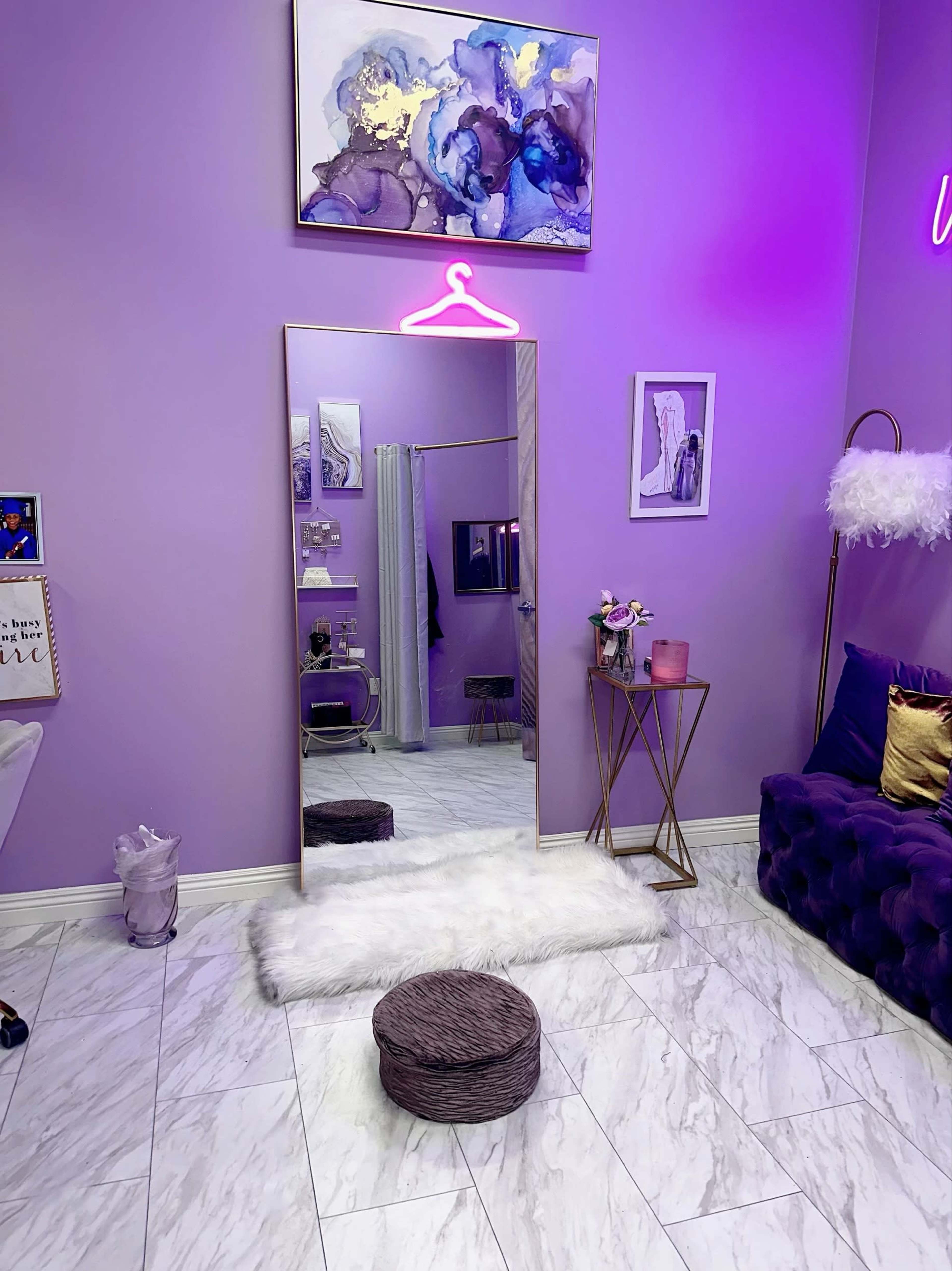 The room features purple walls, a large mirror with a hanging pink neon sign, a plush chair, and a small round ottoman on a white rug.