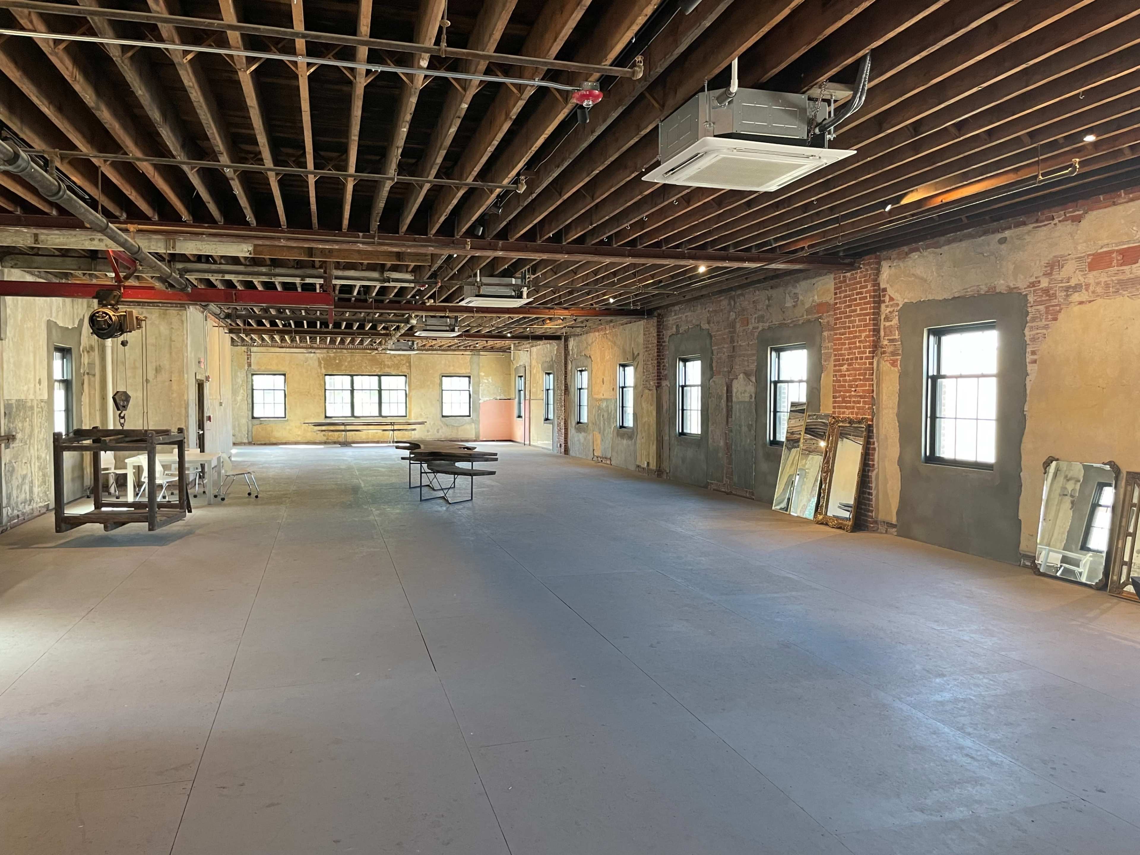 An empty, unfinished room with exposed beams, several windows, and a few pieces of furniture, including a table and mirrors along the walls.