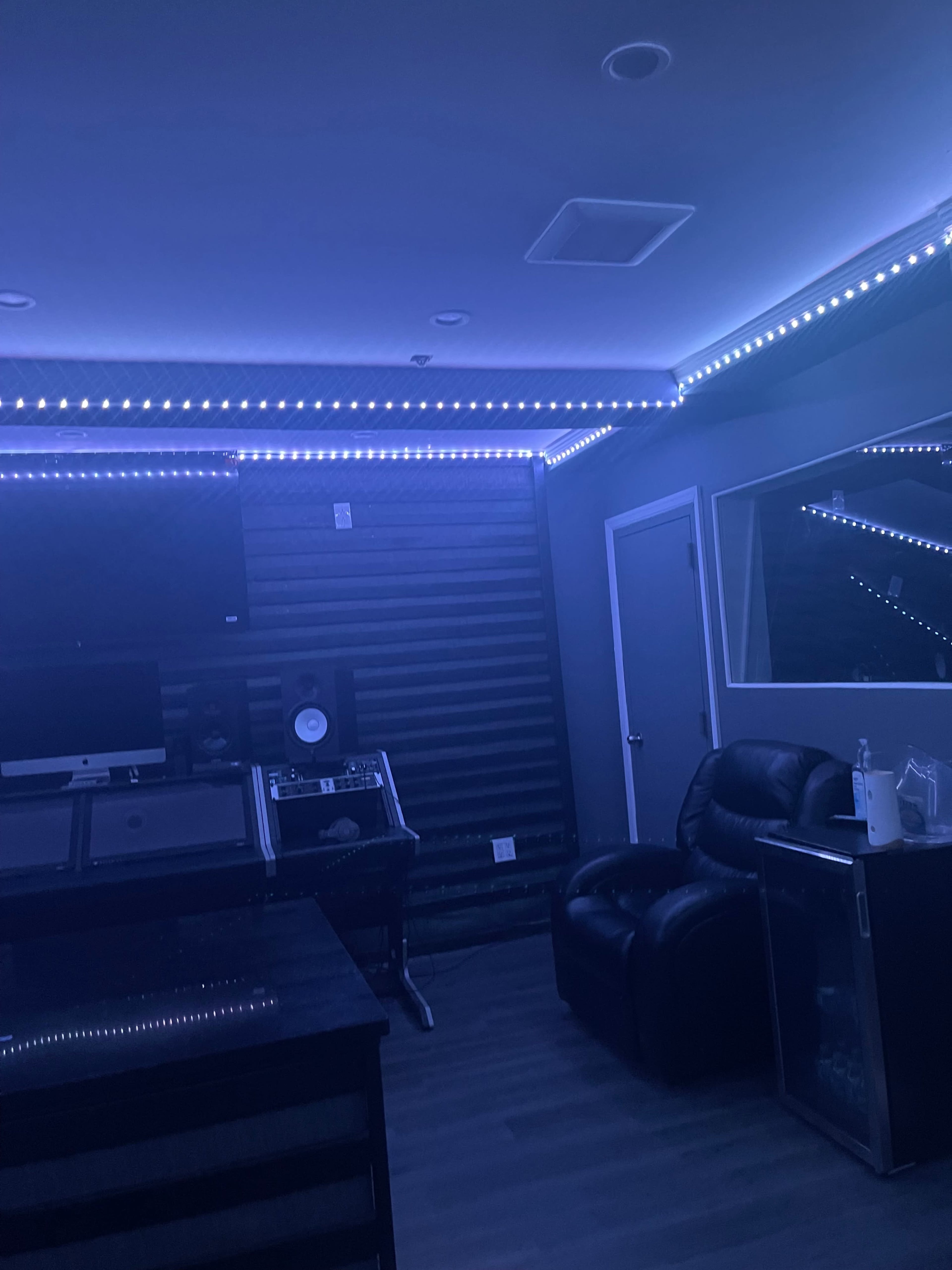 A dimly lit room with blue LED lights, featuring a mixing console, a computer setup, and a black leather chair.