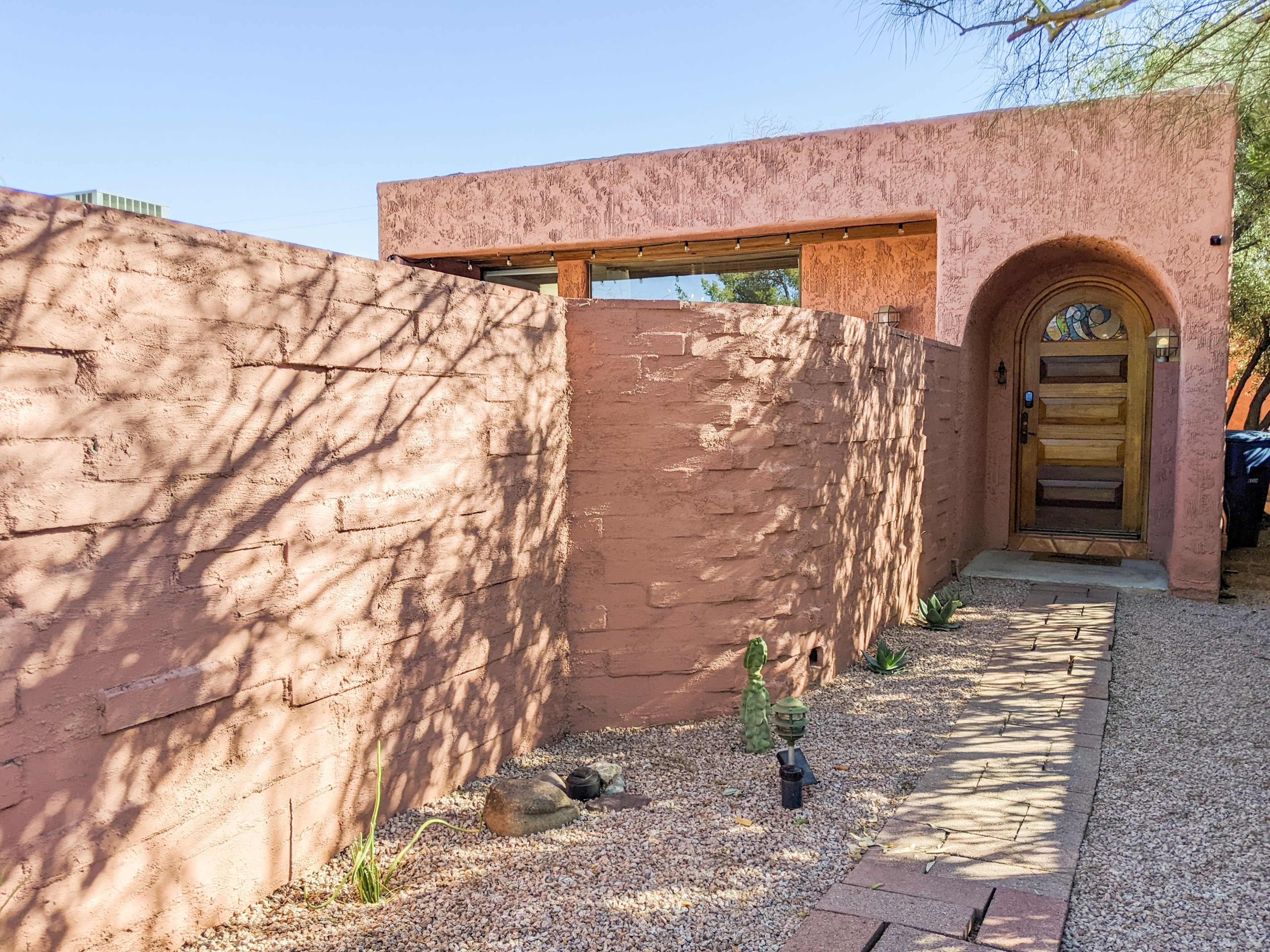 A pink adobe wall surrounds a pathway leading to a wooden door with an arched entrance.
