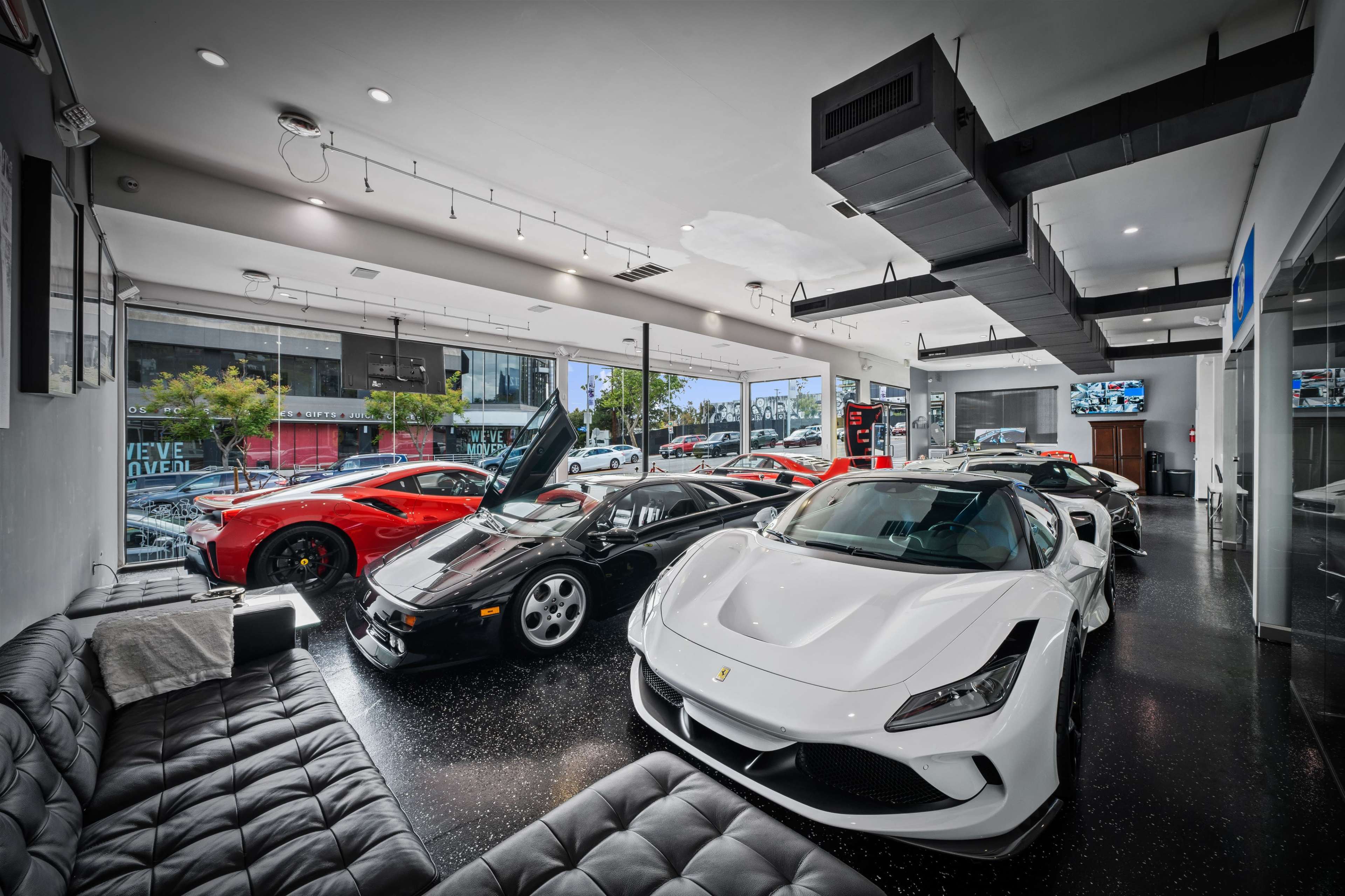 A sleek showroom displays several luxury sports cars, including a white Ferrari, a black Ferrari, and multiple red models, with large windows showcasing the street outside.