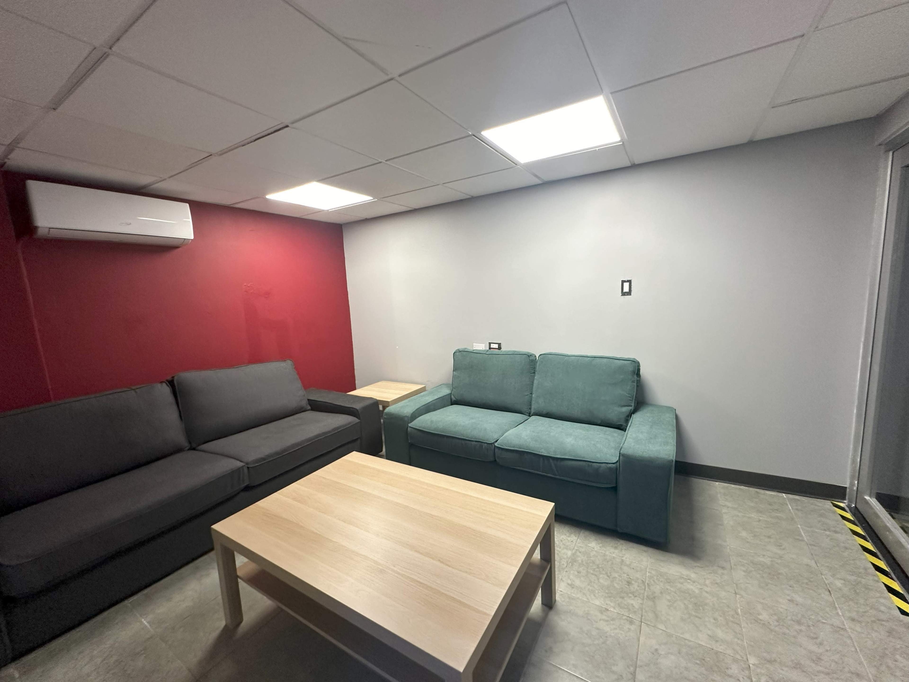 The image shows a small lounge area with two couches, a wooden coffee table, and a light gray wall contrasting with a red feature wall.