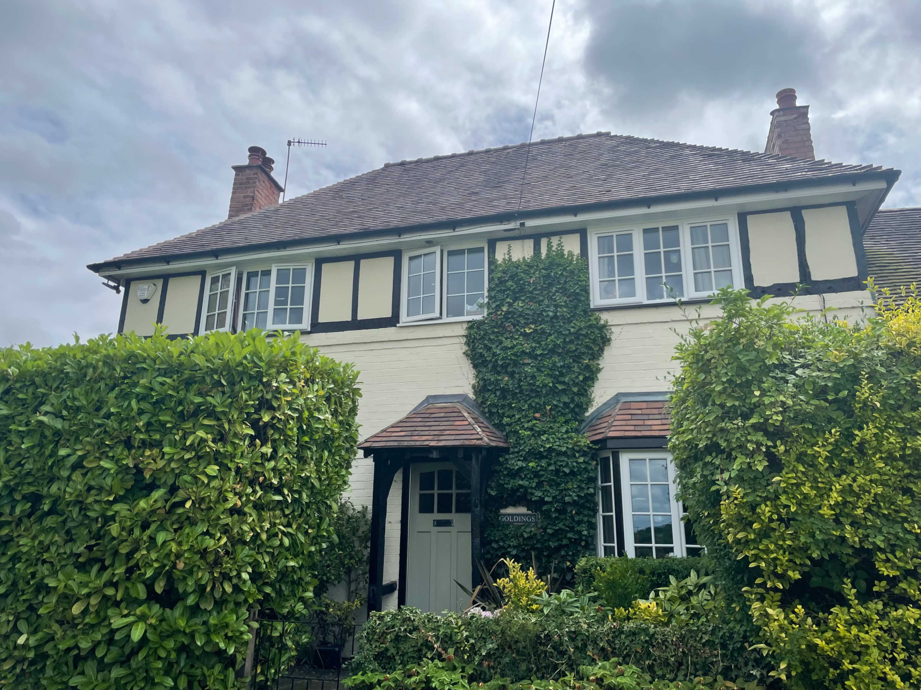 A two-story house with a symmetrical facade is partially obscured by green hedges and climbing vines under a cloudy sky.