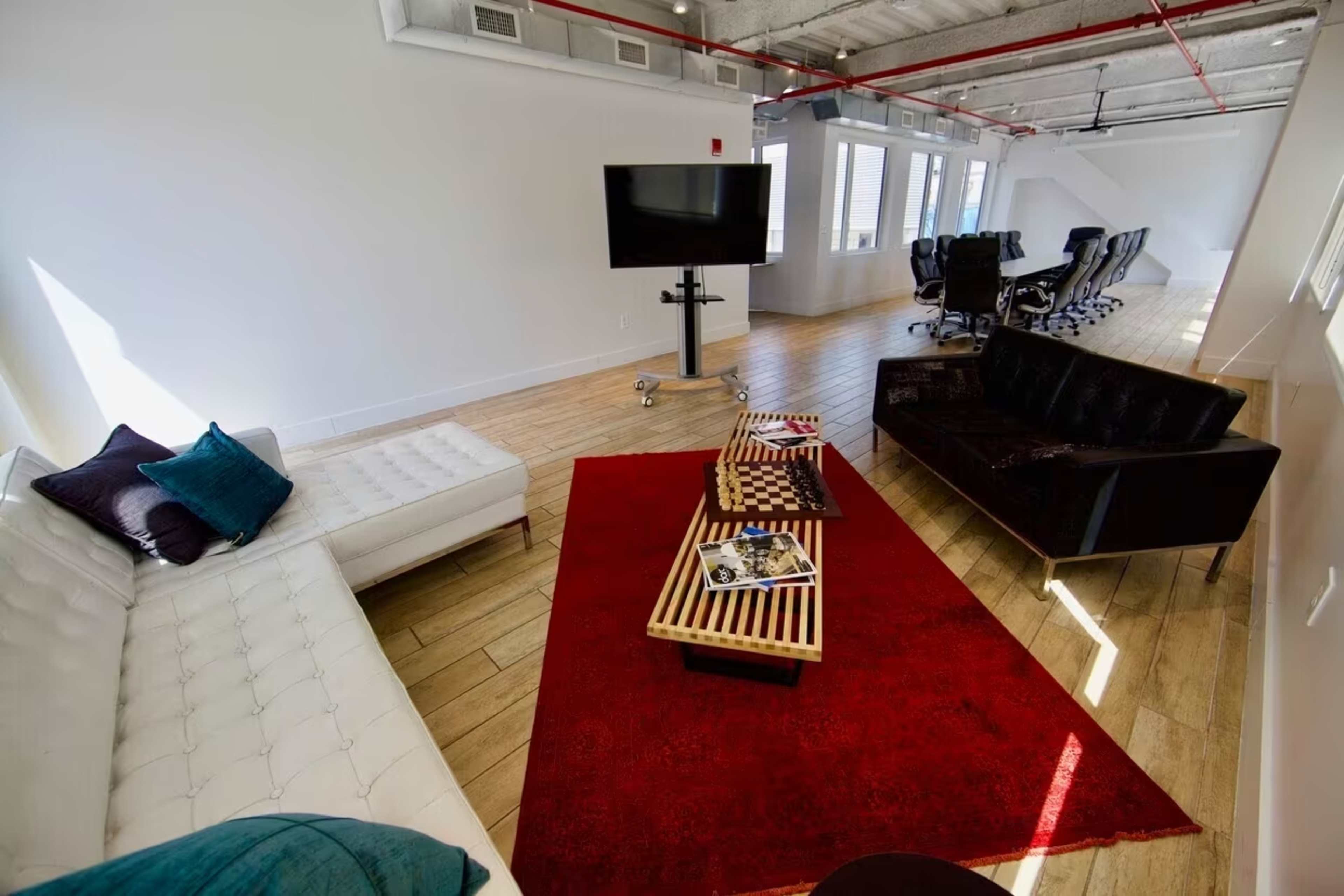 A modern office lounge area featuring a white sofa, a black couch, a red area rug, and a television mounted on a stand.