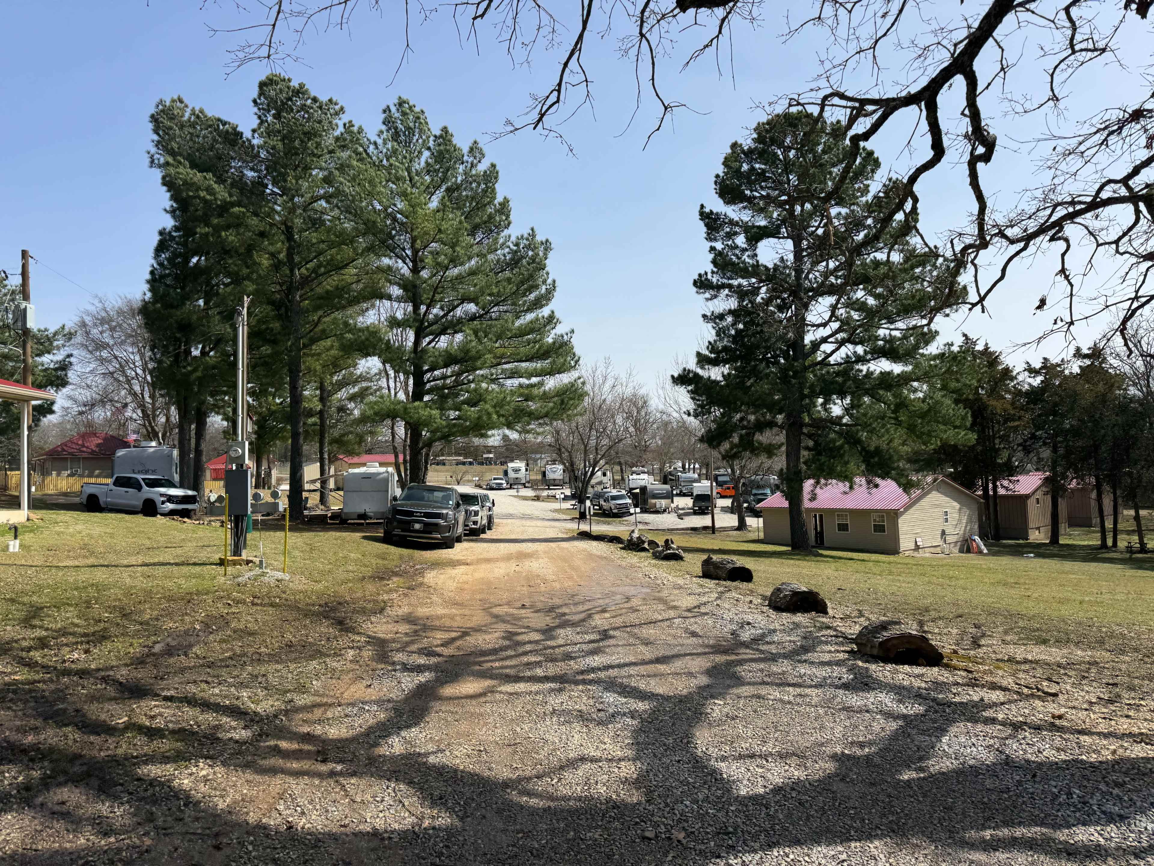 The scene shows a gravel road leading through trees to an area with parked vehicles and small buildings in the background.