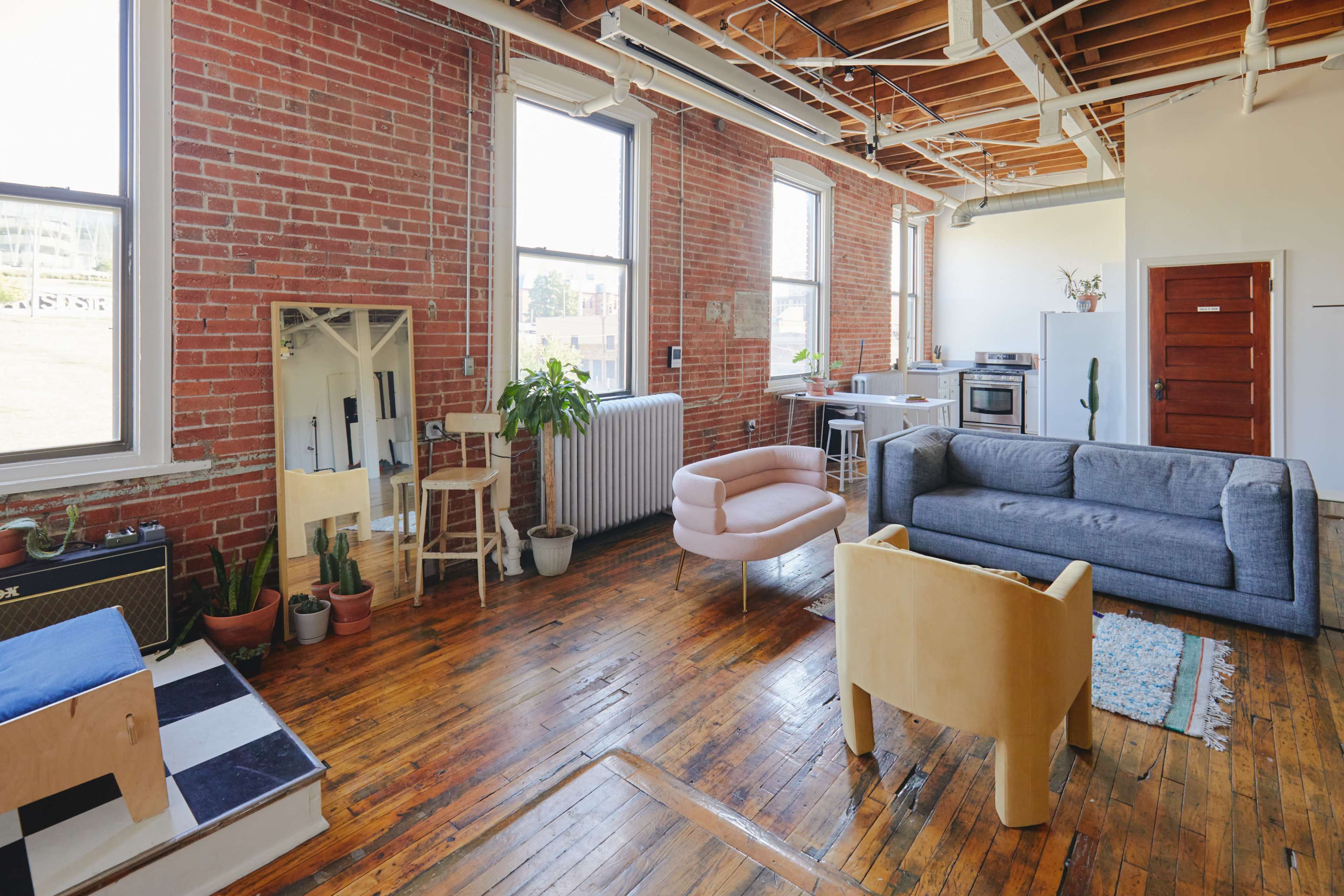 The spacious interior features exposed brick walls, large windows, wooden floors, and a mix of modern furniture, including a pink chair and a gray sofa.