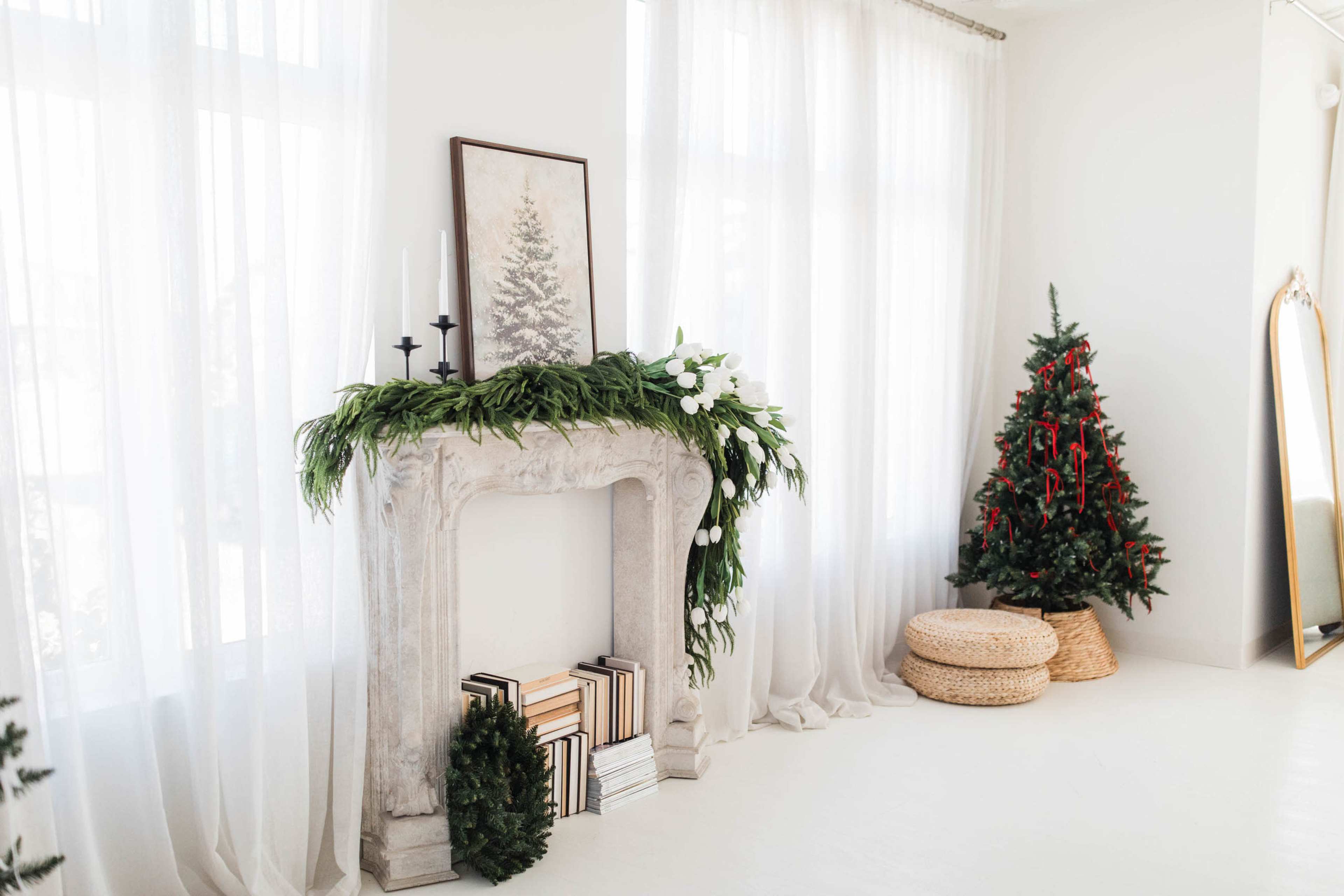 A white room features a decorated fireplace with greenery atop, flanked by a Christmas tree and a mirror.