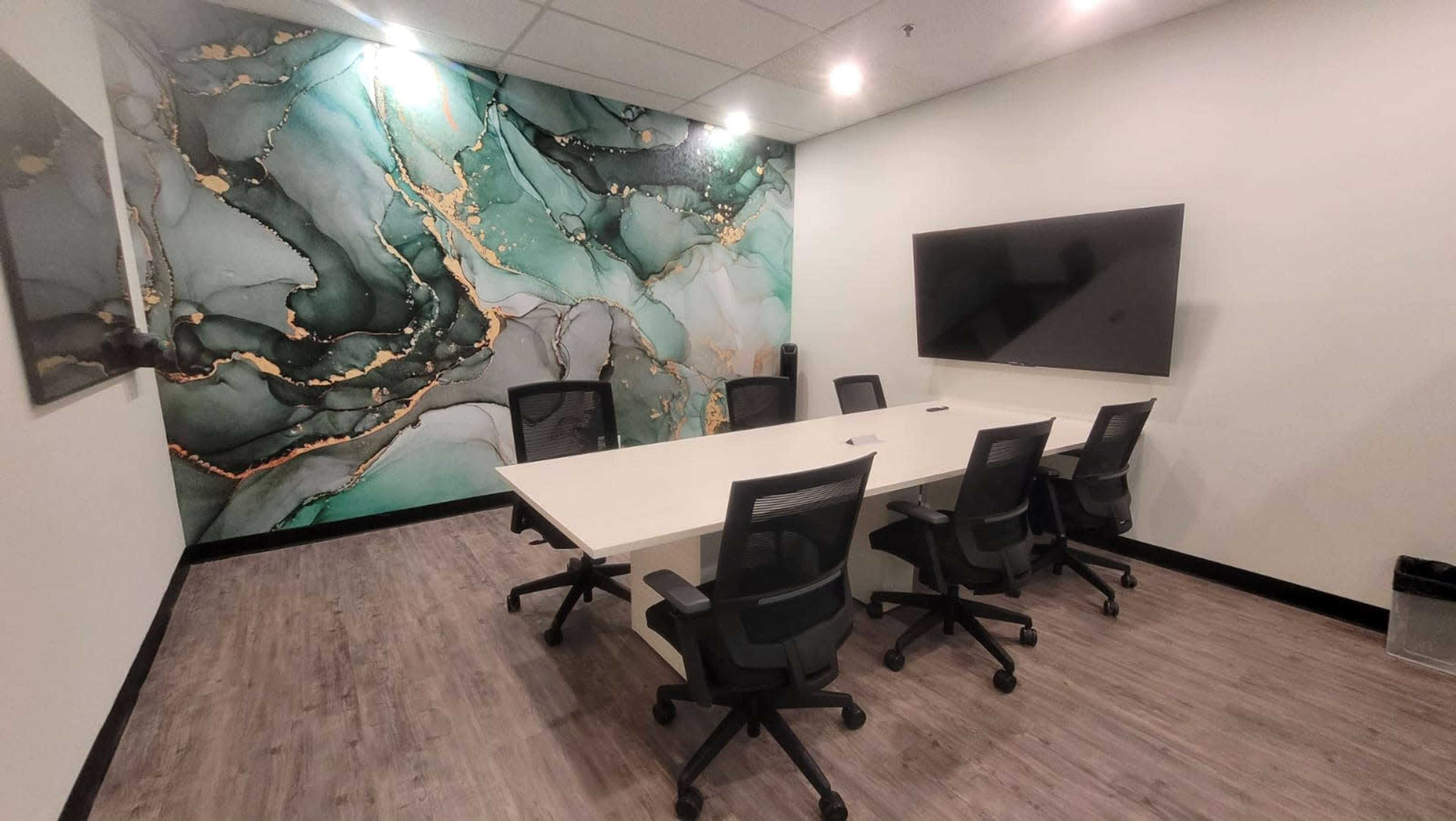 A modern conference room features a large rectangular table surrounded by six black office chairs, with an abstract green and gold wall mural and a wall-mounted TV.