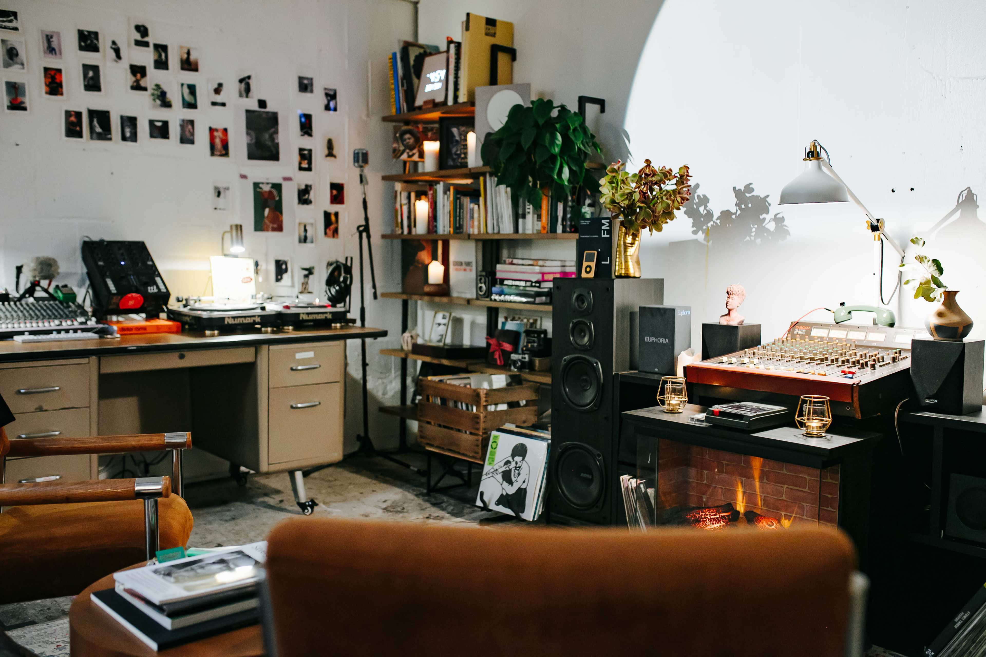 A cozy music studio features a desk with recording equipment, a wall adorned with photos, a bookshelf filled with books, and a small fireplace, all surrounded by comfortable seating.