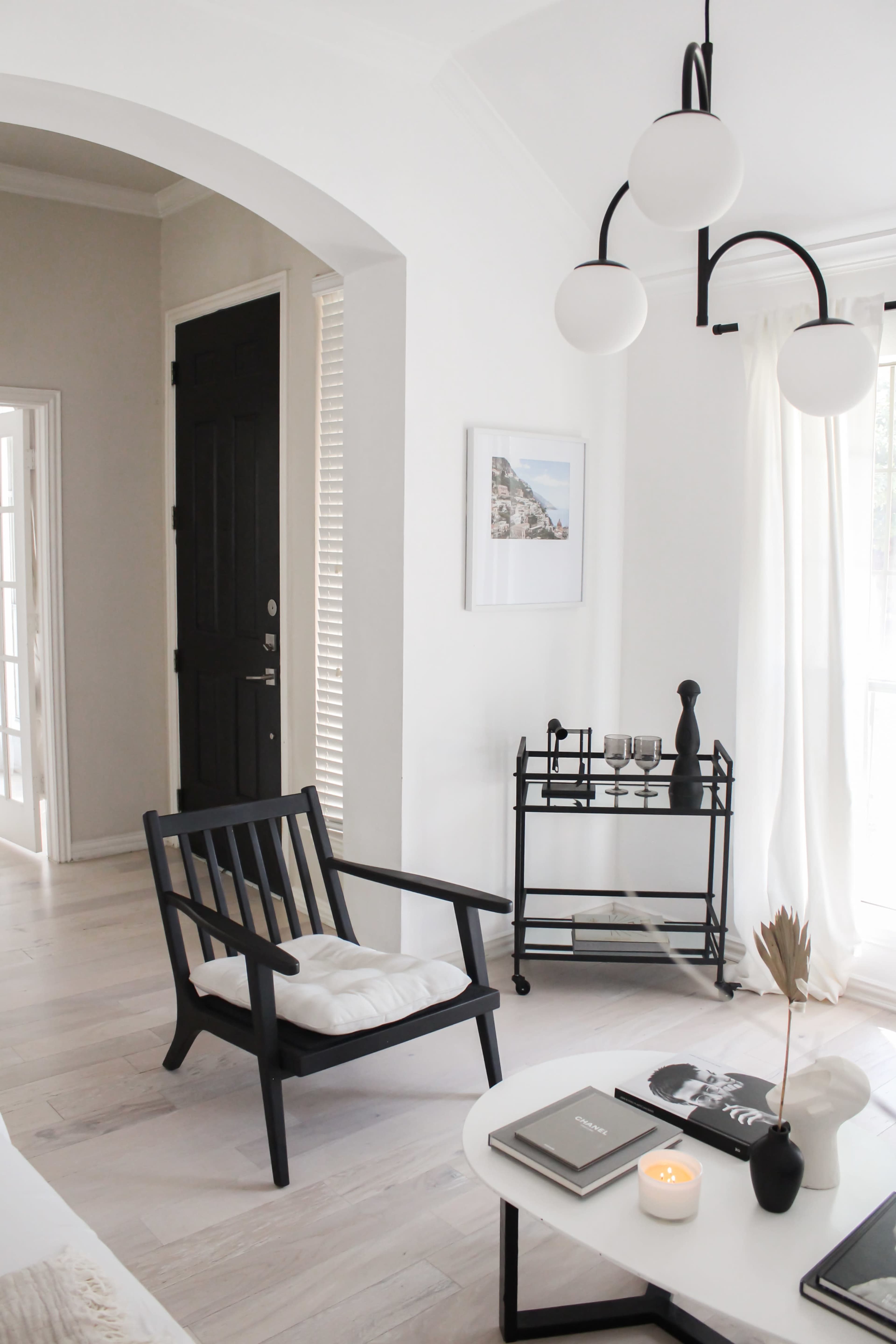 The image shows a minimalist living space featuring a black wooden chair, a round coffee table with books and a candle, and a small bar cart beside a large window.