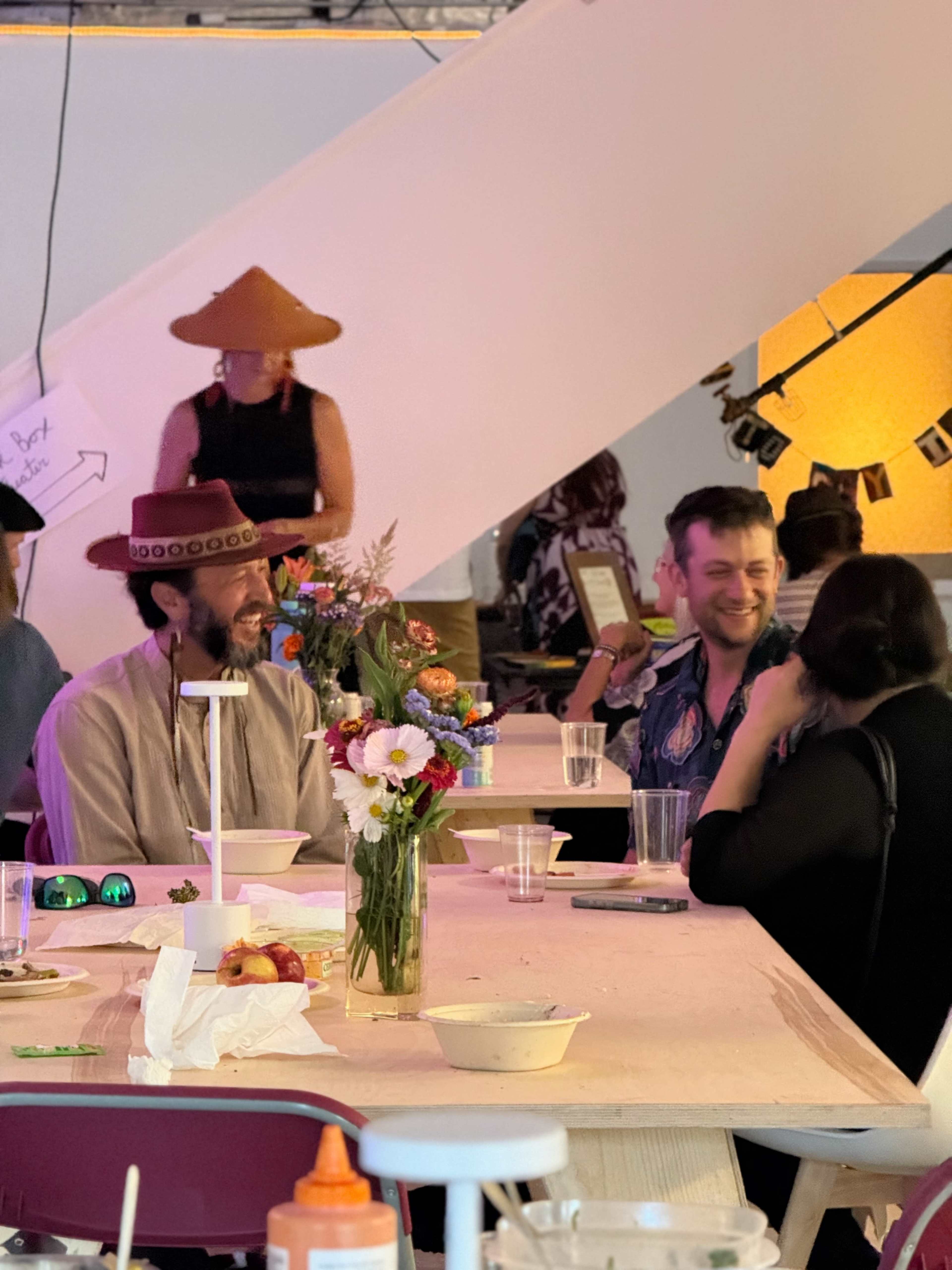 The image depicts a group of people sitting around a table adorned with flowers and food, while one person in the background wears a large, distinctive hat.