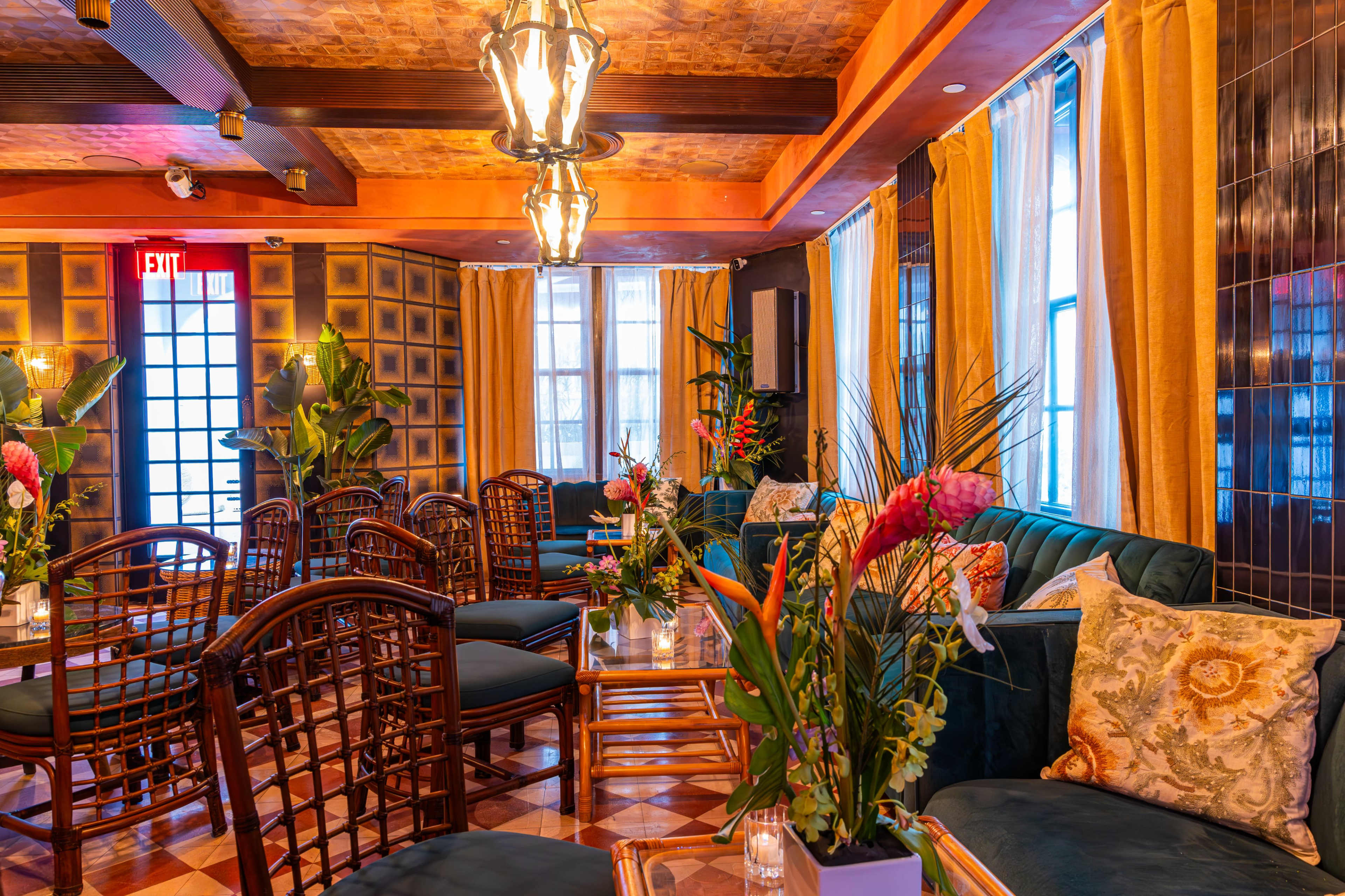 The image shows a stylish lounge area with green sofas, wooden furniture, and abundant floral arrangements, illuminated by hanging lights.