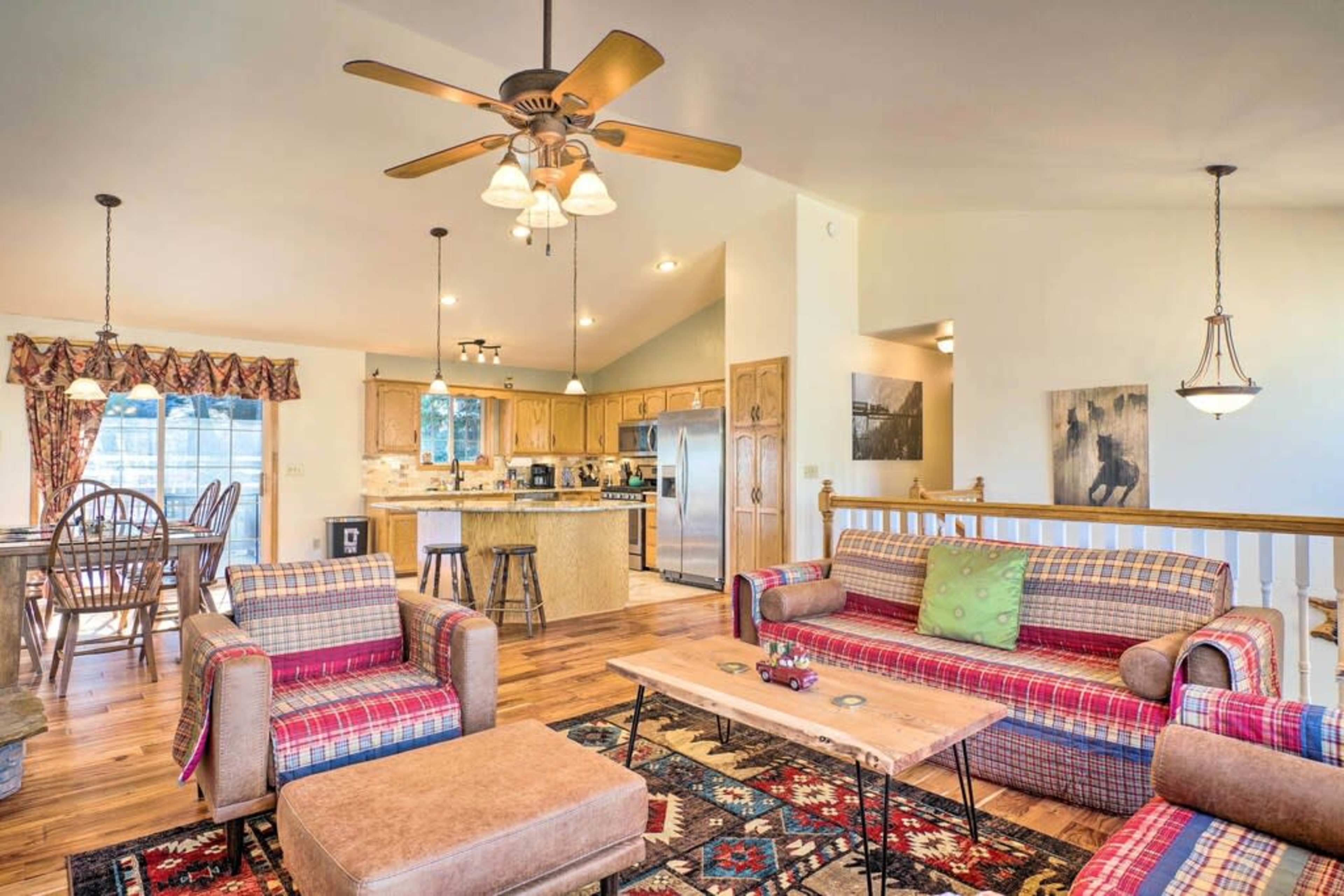 The image shows a cozy living room with patterned couches, a wooden coffee table, and an open kitchen area in a well-lit, spacious setting.