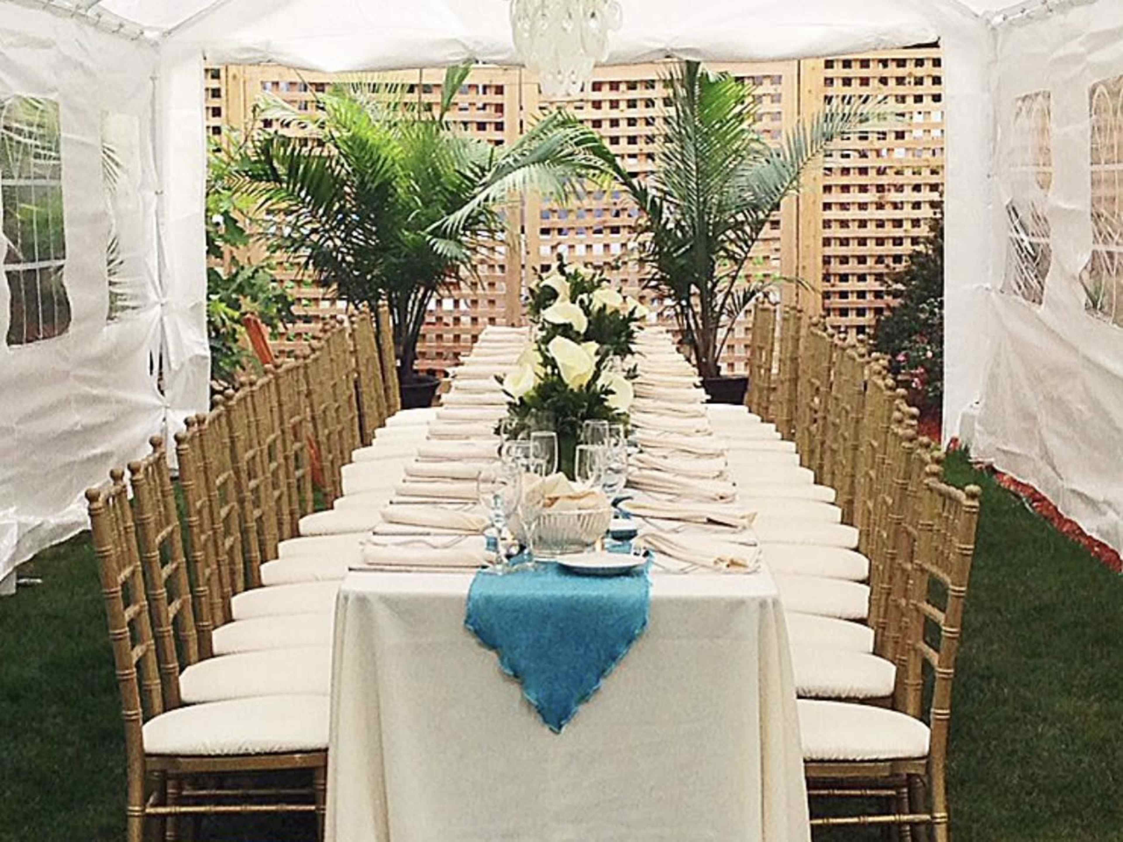 A long dining table is set up under a white tent, surrounded by chairs and decorated with a centerpiece of flowers and palm plants.