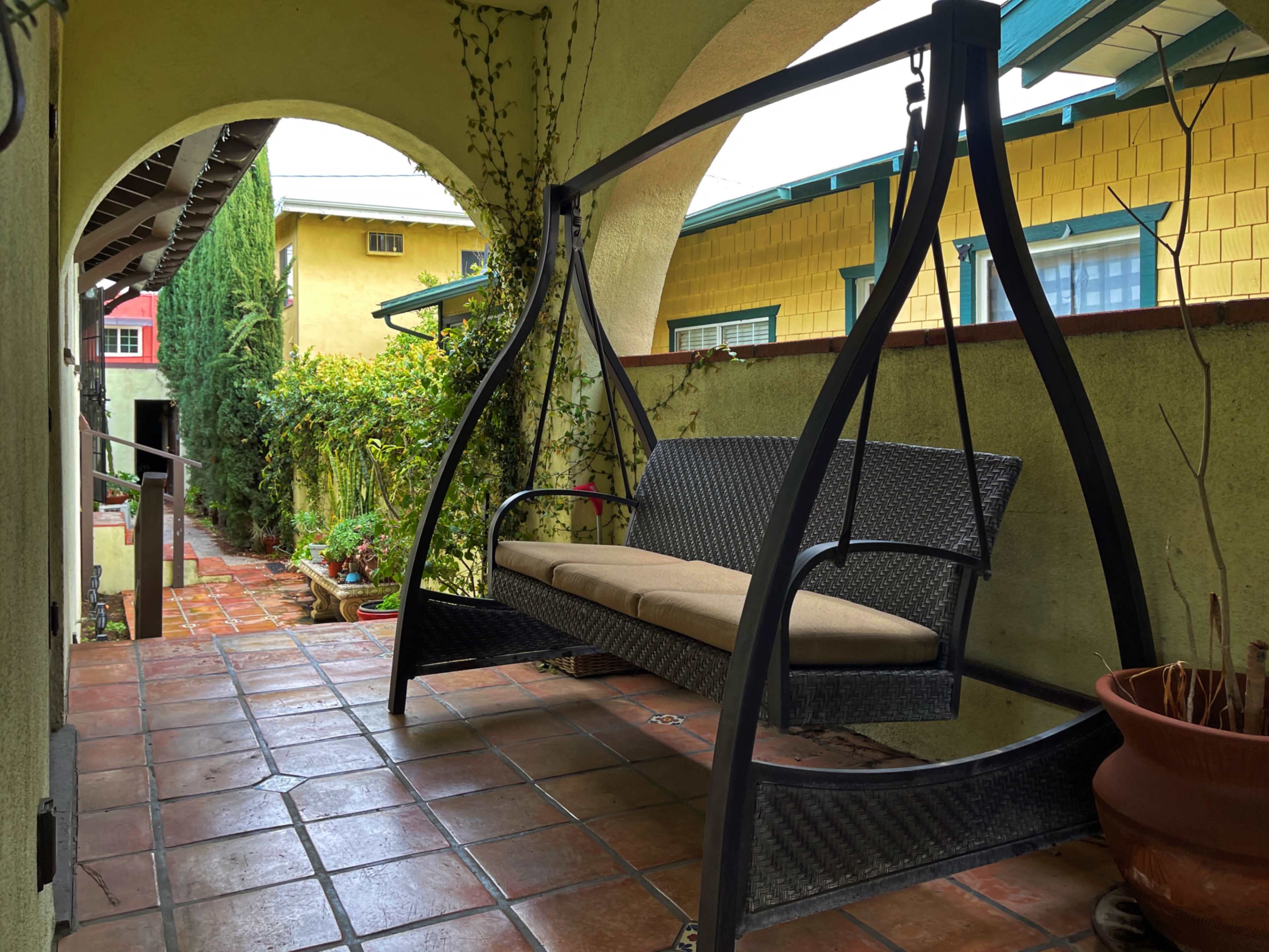 A black swing seat is positioned under an archway, flanked by tiled pathways and greenery on either side.