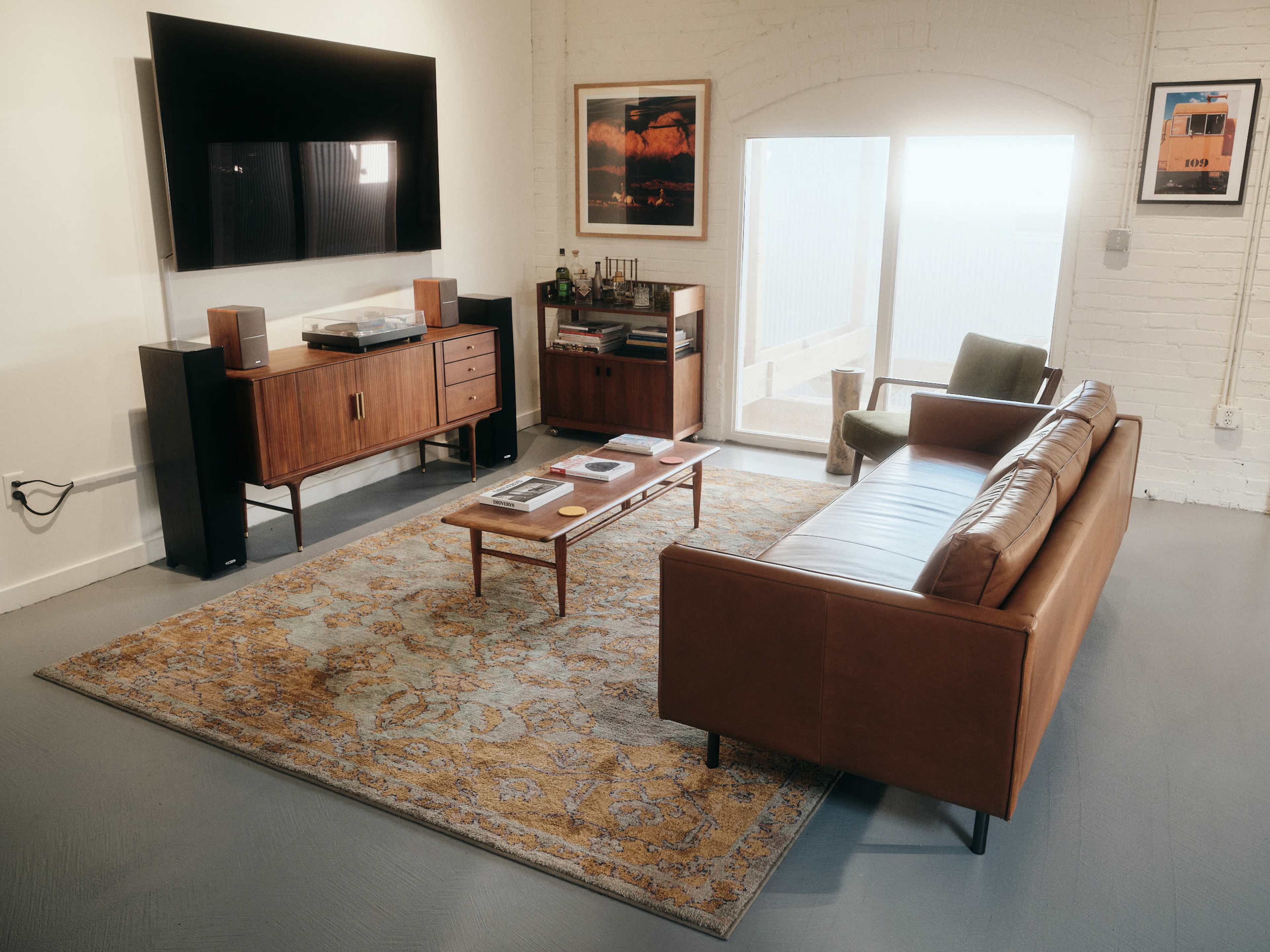 A modern living room features a leather sofa, wooden furniture, a television mounted on the wall, and a patterned rug with a coffee table in the center.