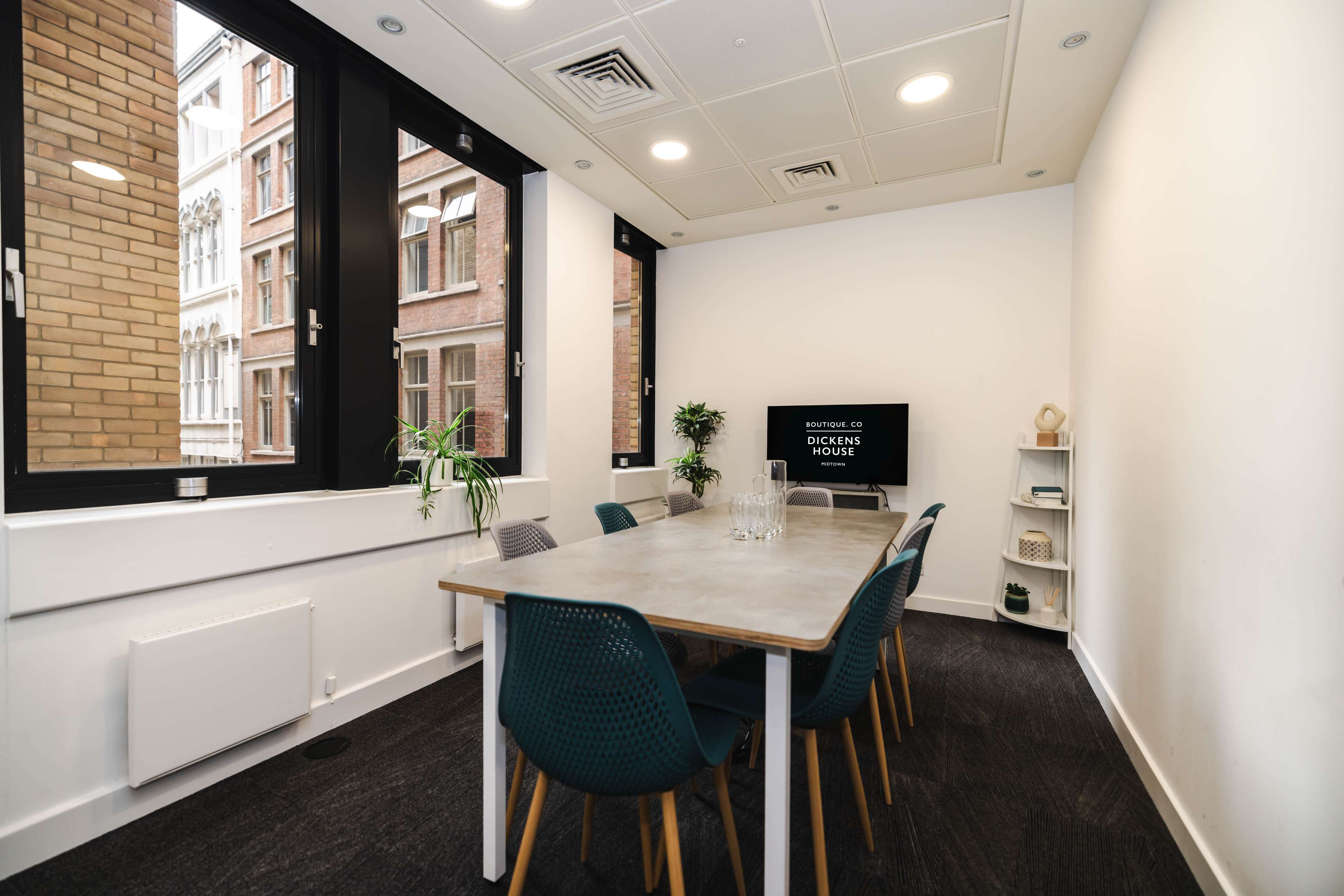 A modern meeting room with a long table surrounded by chairs, large windows providing natural light, and a television mounted on the wall.