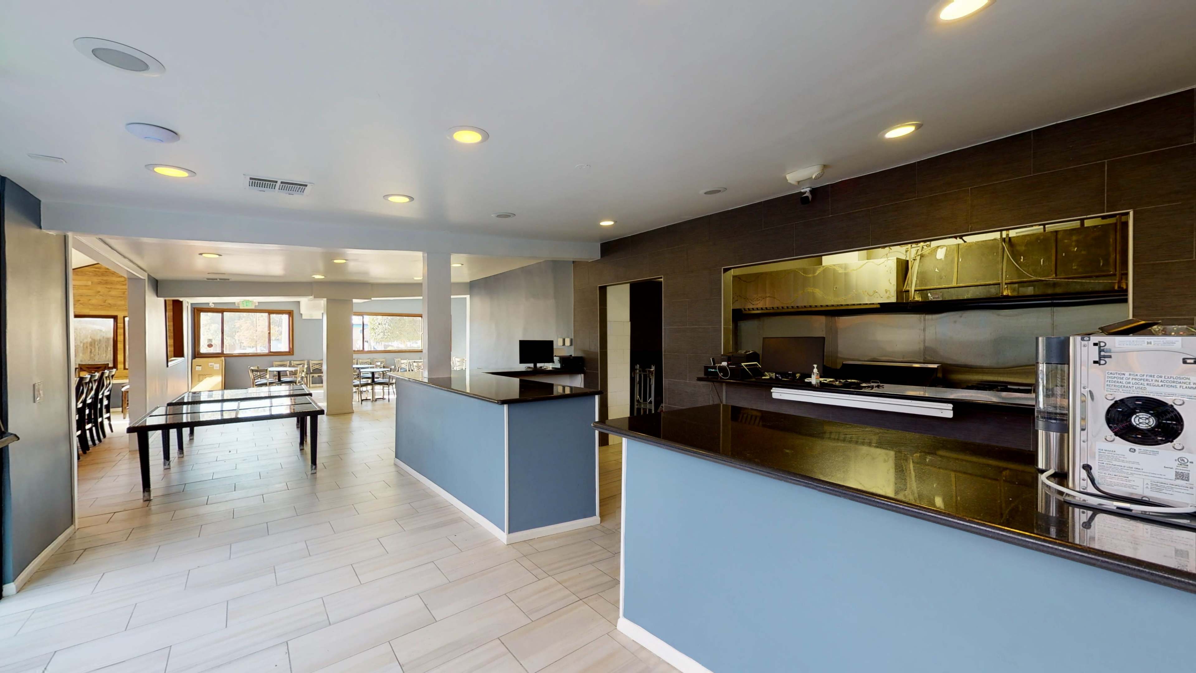 The image shows a modern kitchen area with dark countertops and a stainless steel cooking range, adjacent to a dining space with large windows and wooden tables.