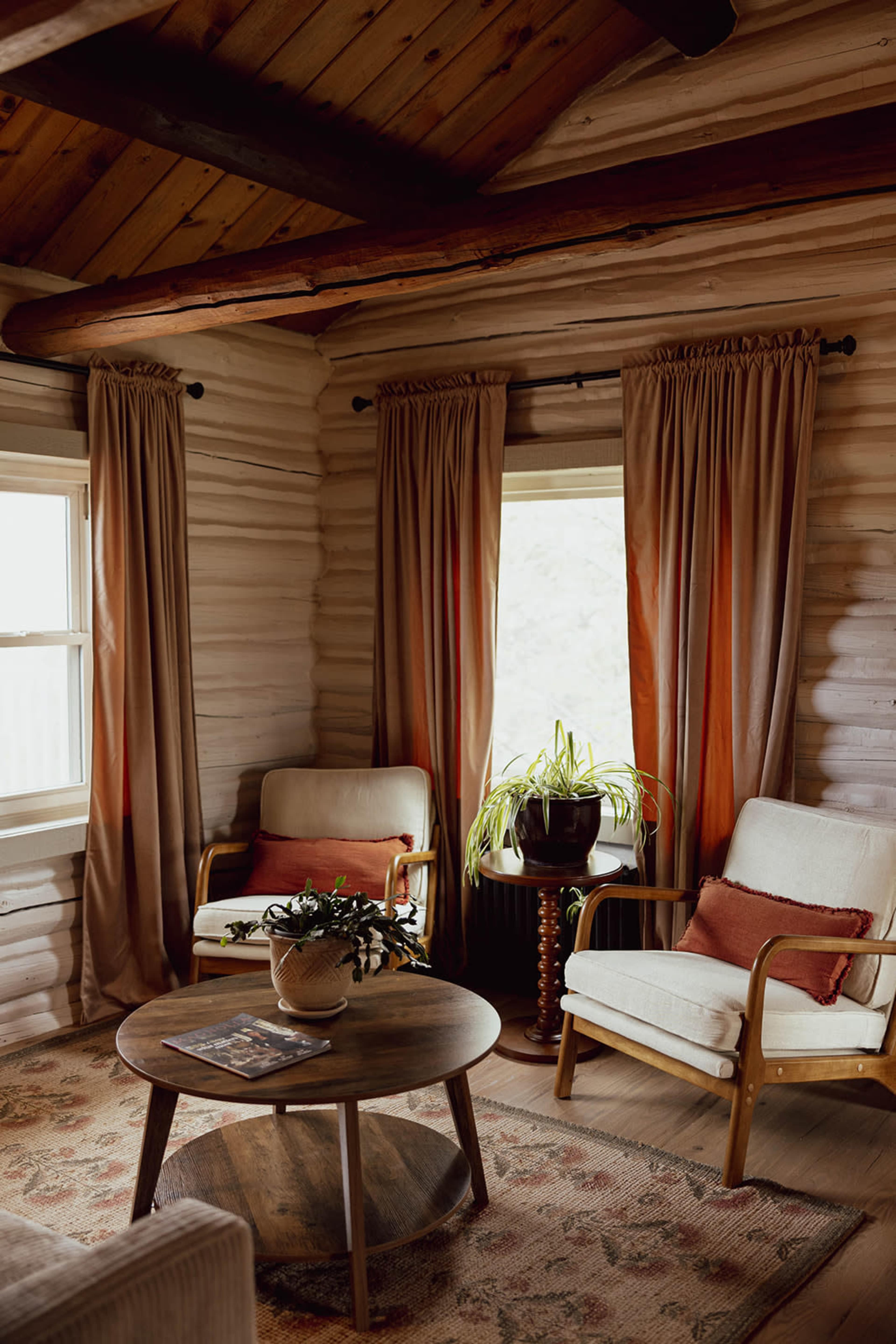 The image shows a cozy living room featuring two upholstered chairs, a round wooden coffee table, and large windows dressed with curtains.