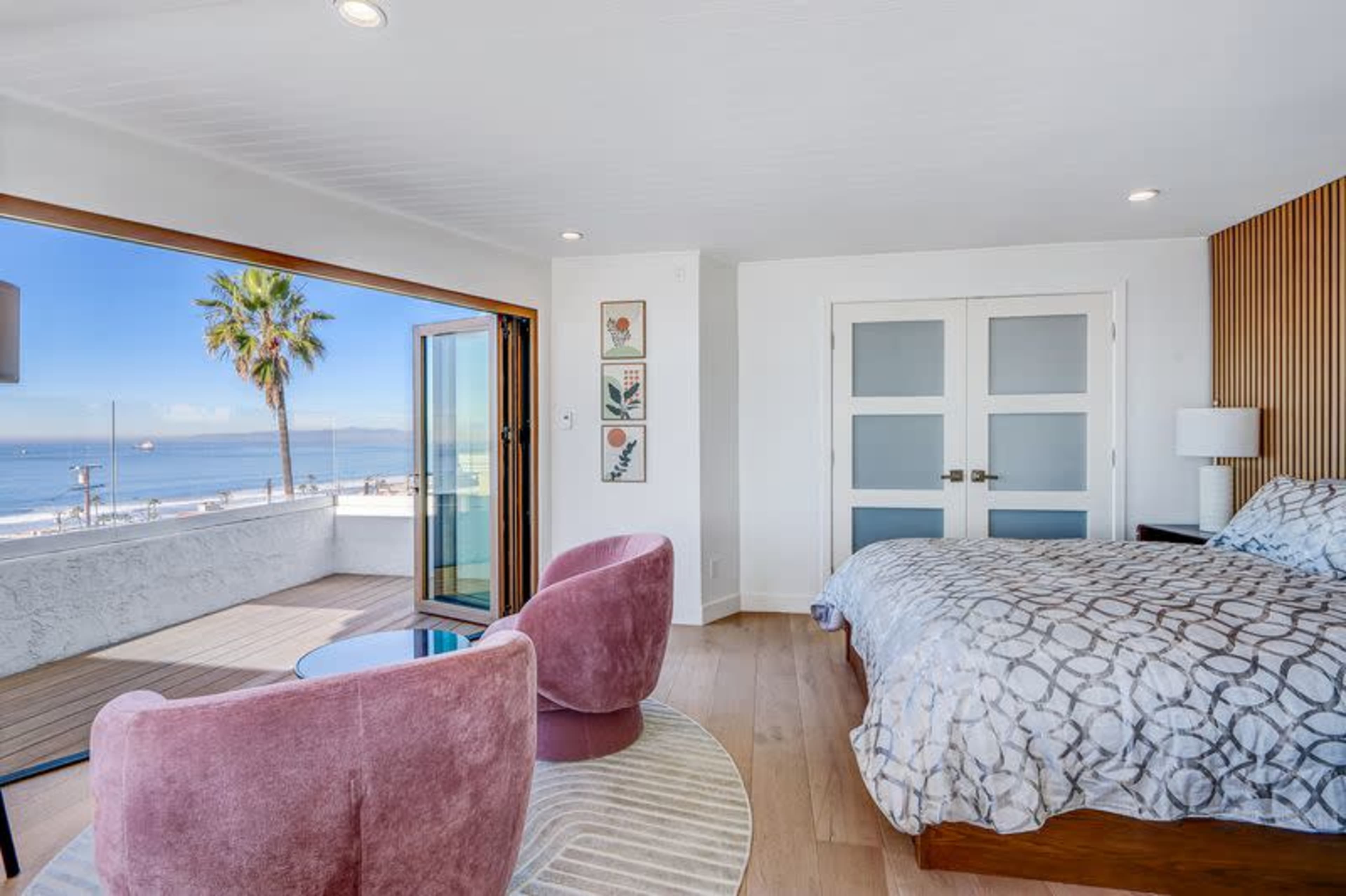 The image shows a modern bedroom with a large window overlooking a beach, featuring a bed, two pink chairs, and wooden accents.