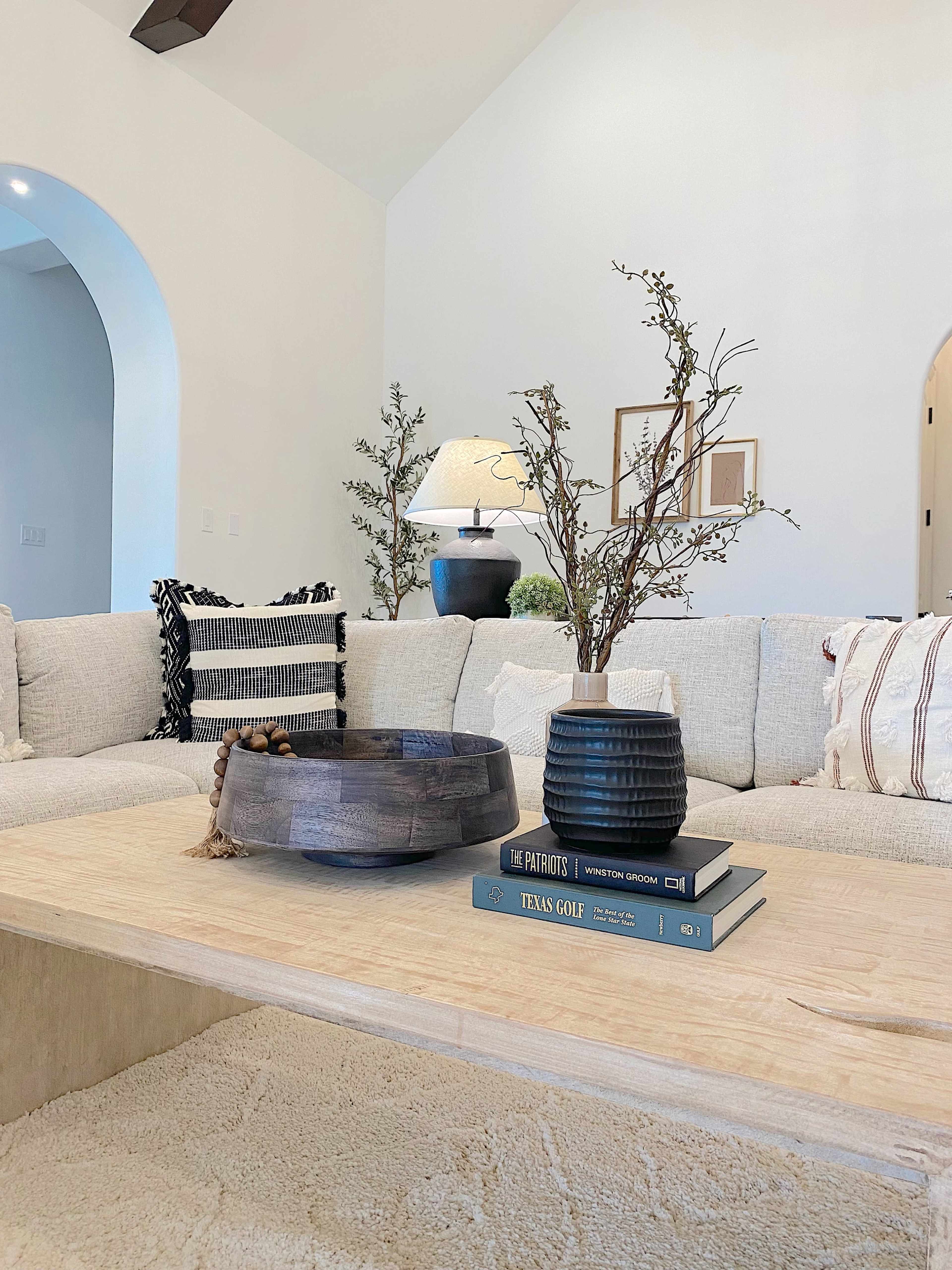 A cozy living room features a light-colored couch with decorative pillows, a wooden coffee table with stacked books and a vase, and a lamp in the background.