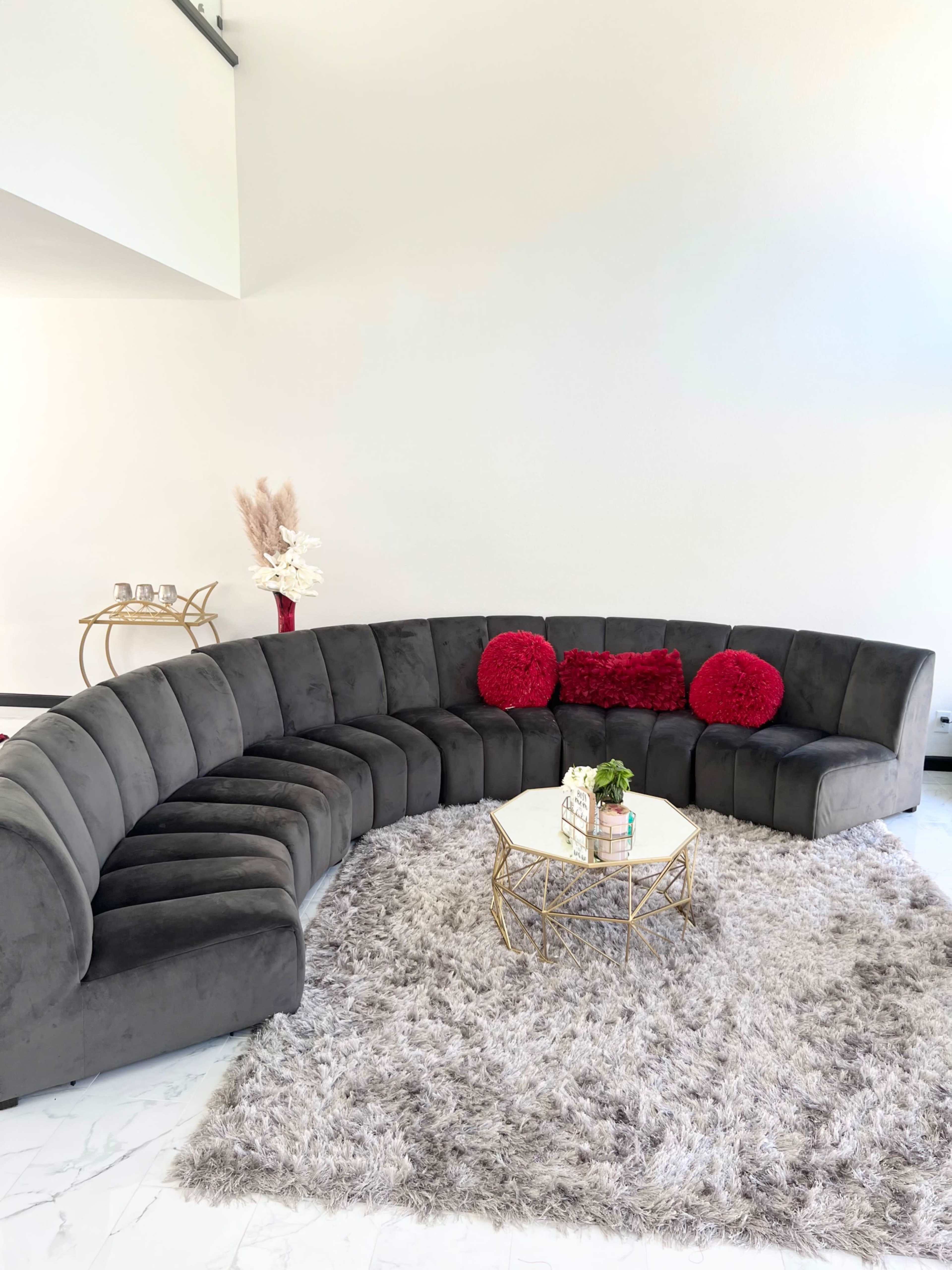 A curved gray sofa with red pillows surrounds a gold coffee table on a plush gray area rug in a bright room.