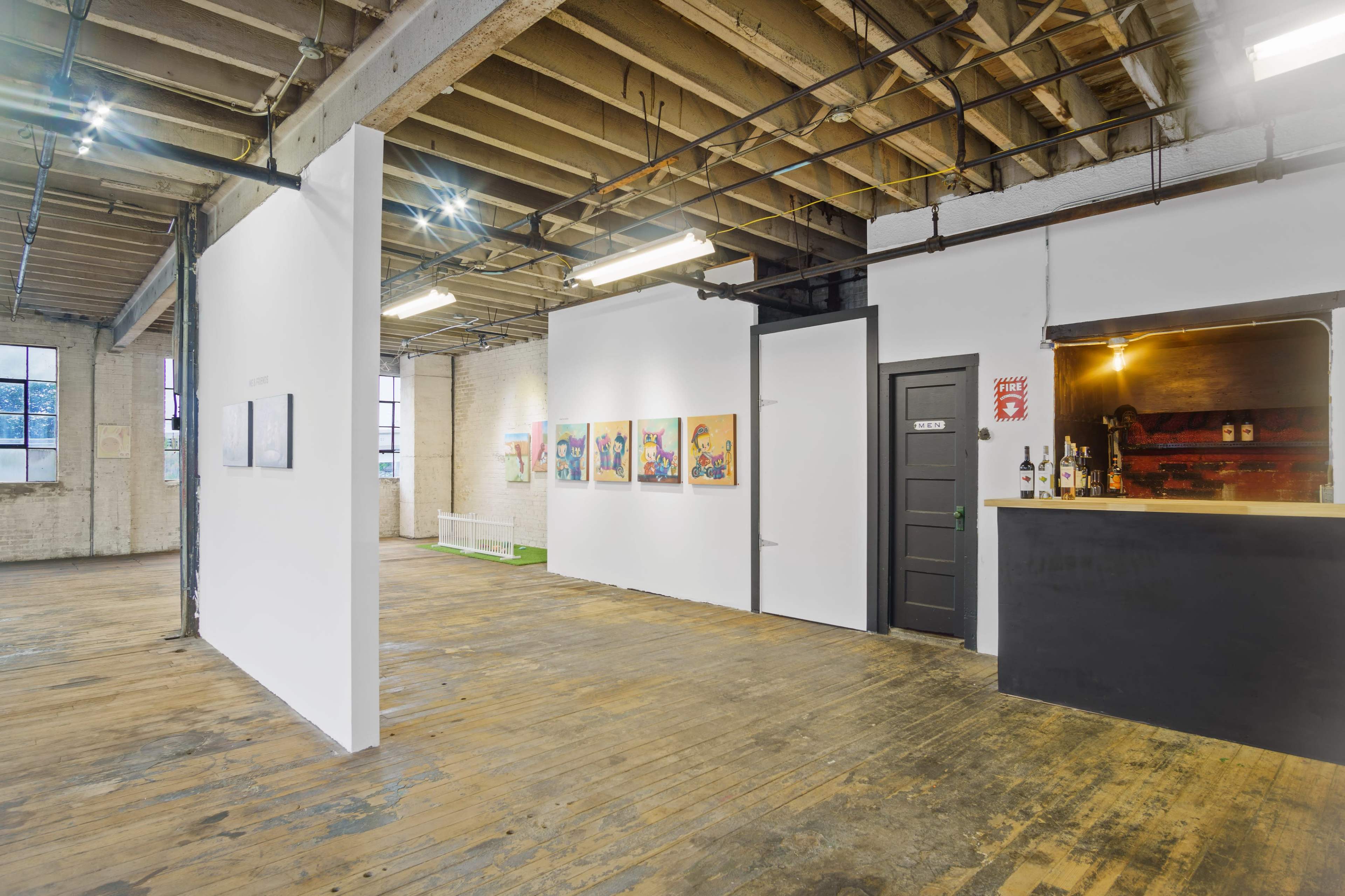 A spacious interior with exposed wooden beams, featuring an art display area with framed artworks on one wall and a bar counter against another.