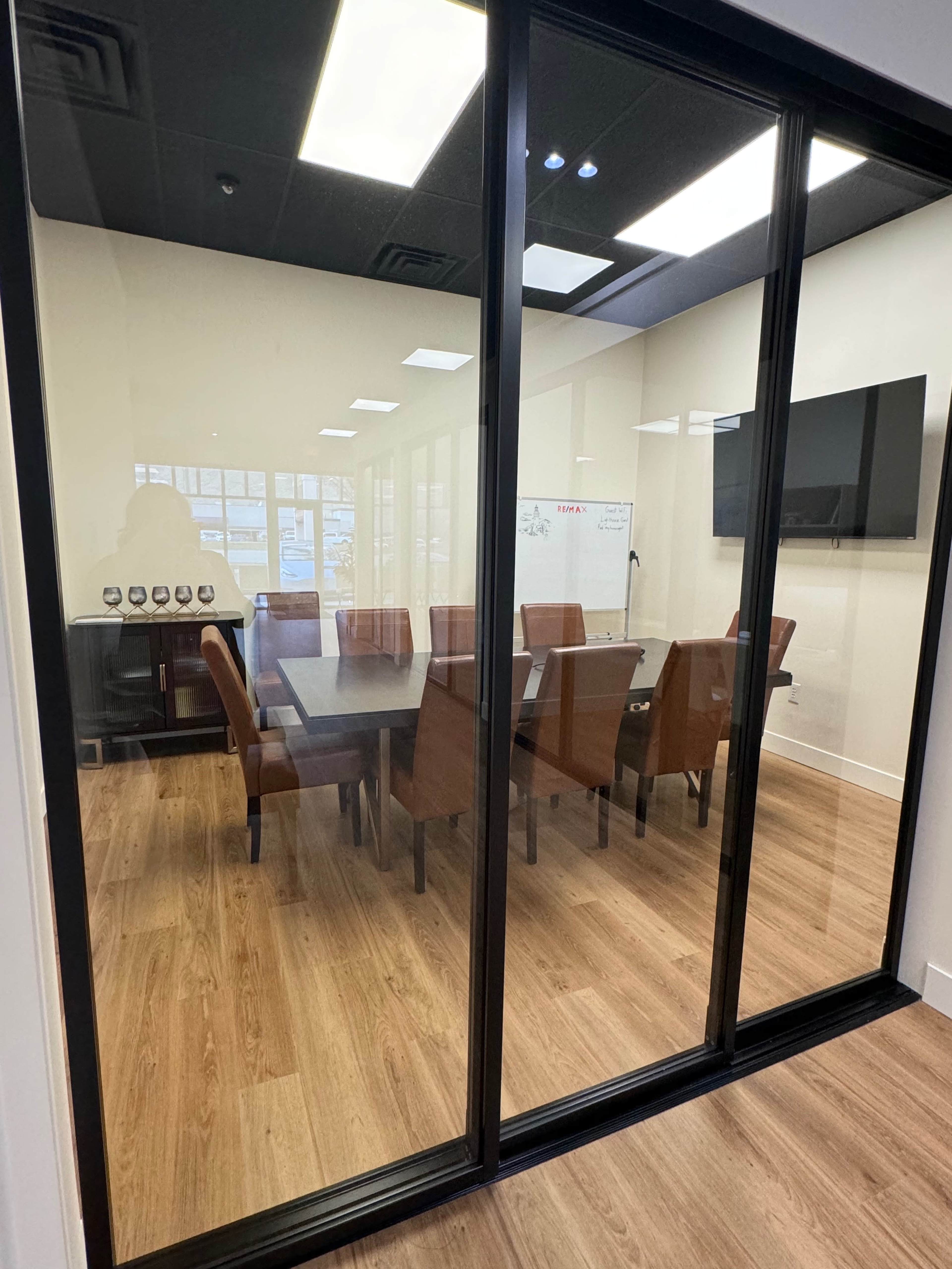 A glass-walled conference room features a large wooden table surrounded by brown leather chairs and a wall-mounted television.