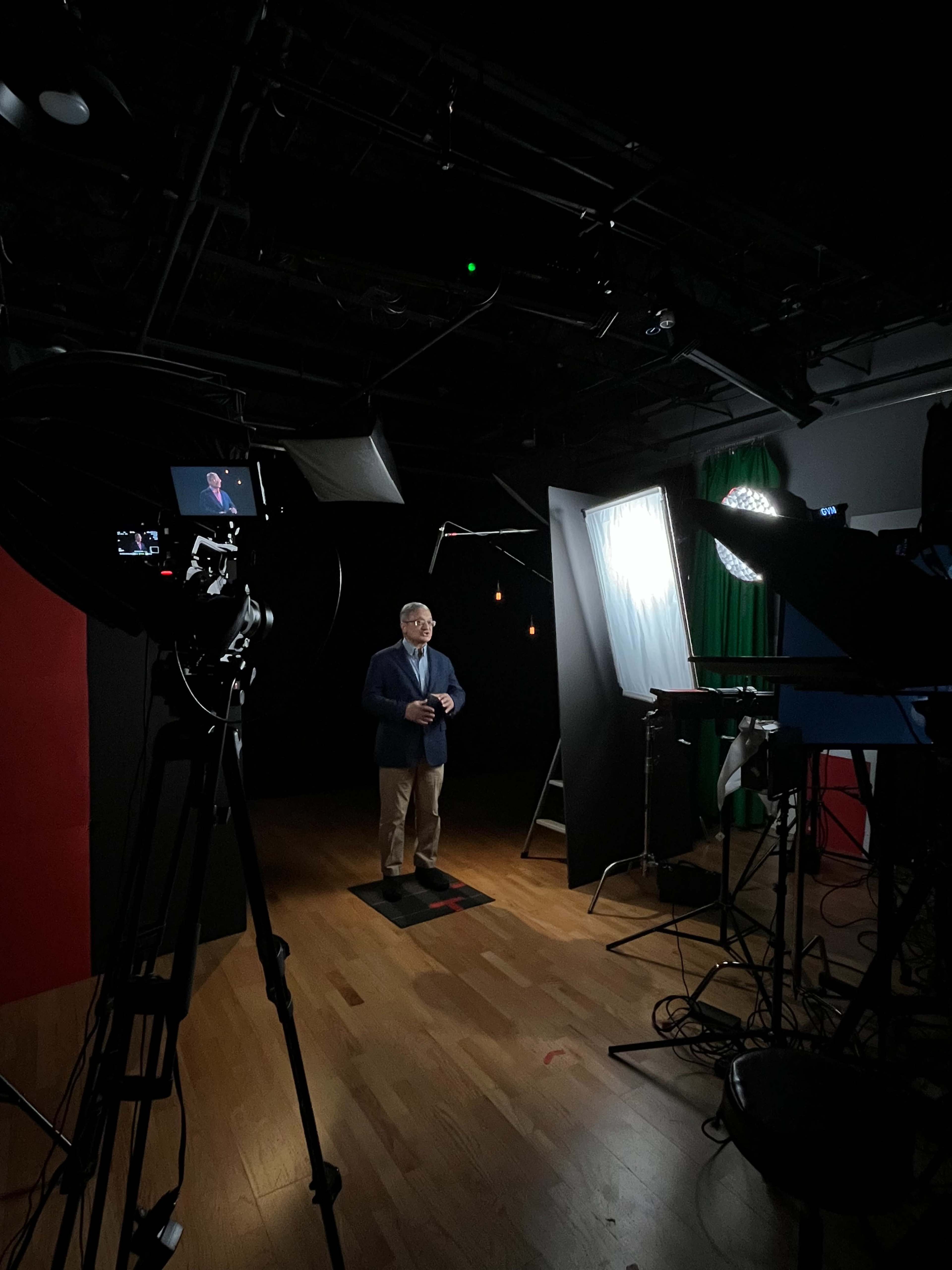 A man stands on a marked spot in a dimly lit studio, surrounded by various lighting equipment and cameras.