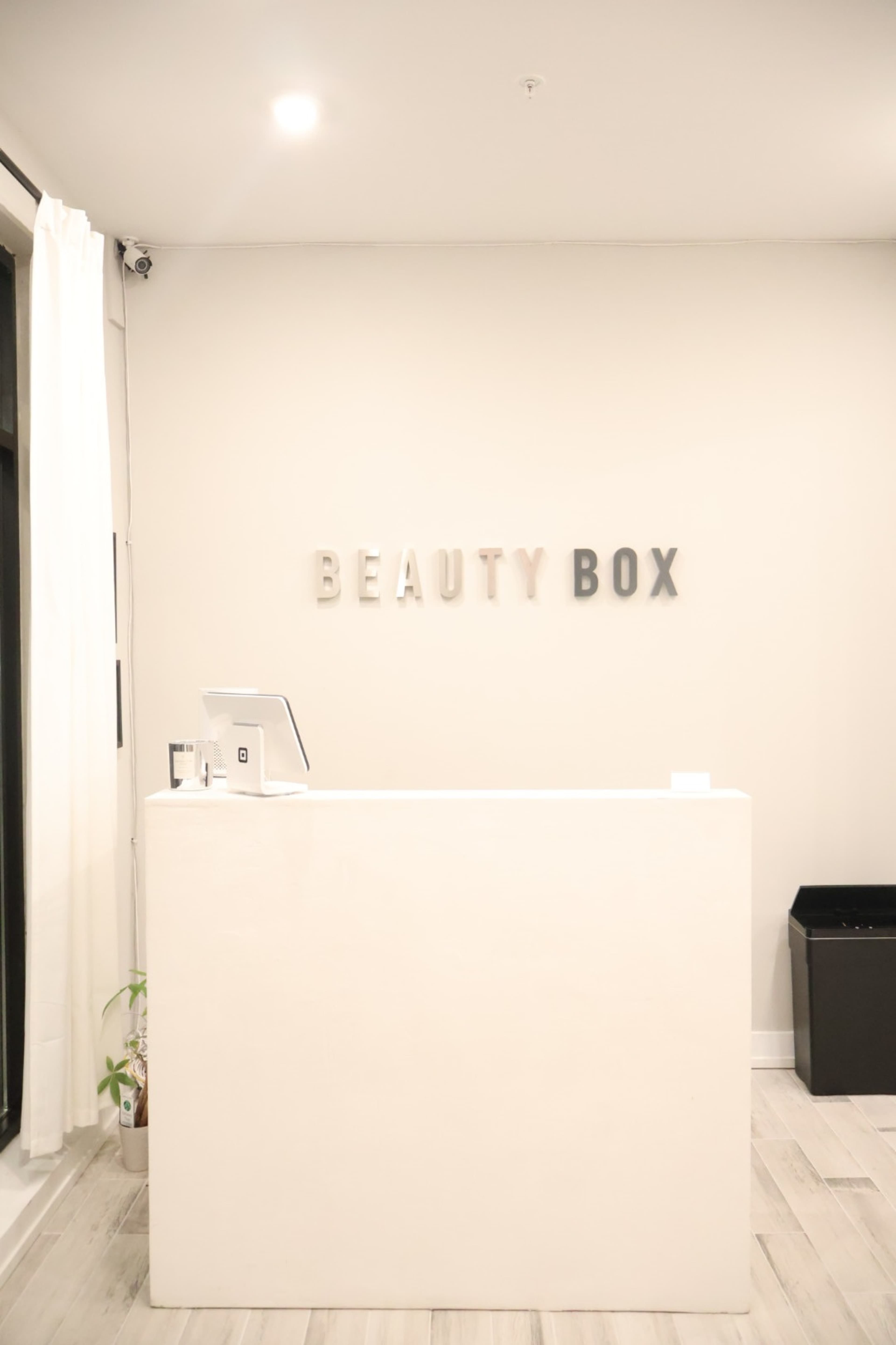 The image shows a minimalist reception area with a white desk and the words "BEAUTY BOX" displayed on the wall behind it.