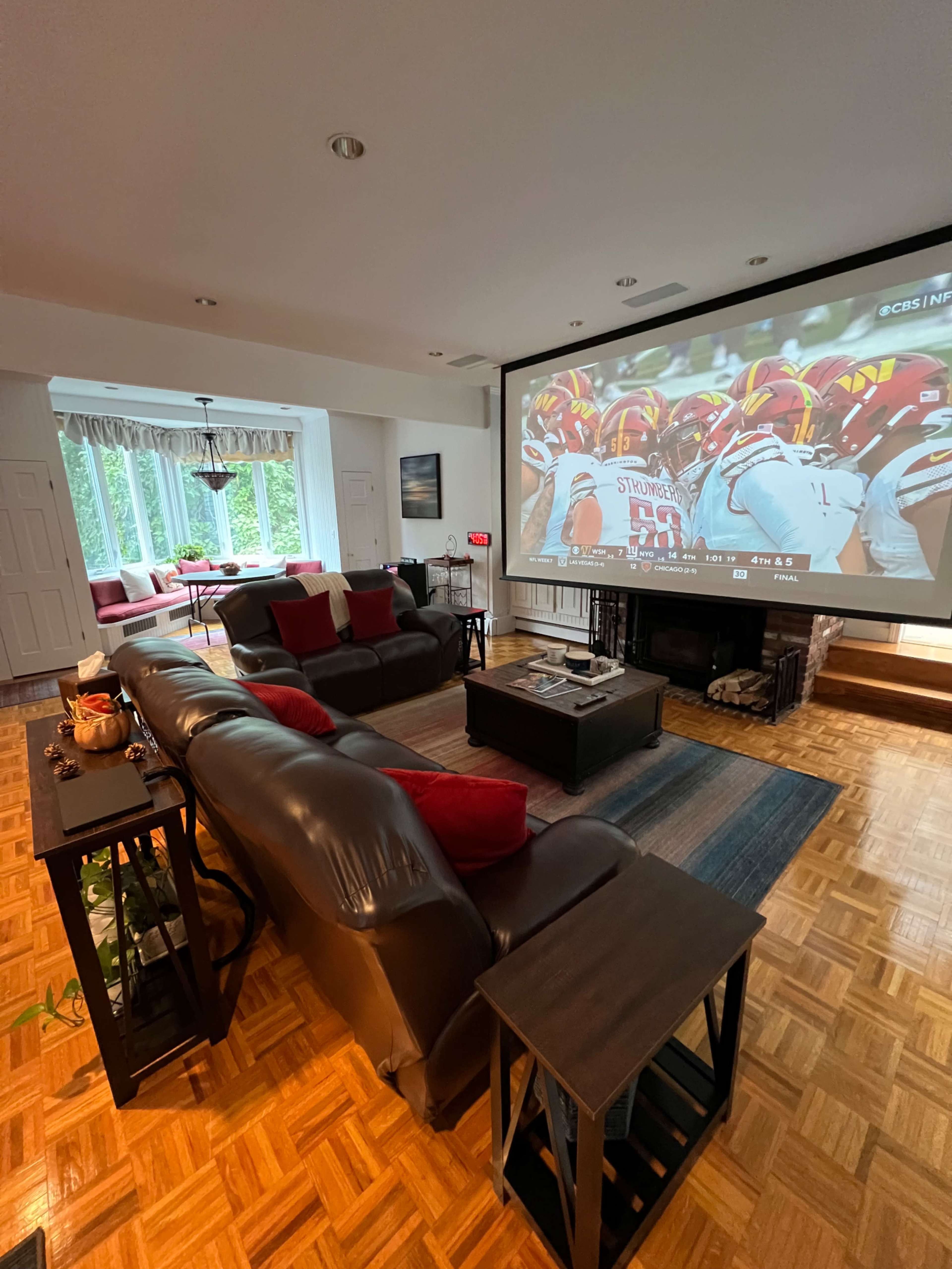 The living room features a large projector screen displaying a sports game, with dark leather sofas and wooden coffee tables arranged on a parquet floor.