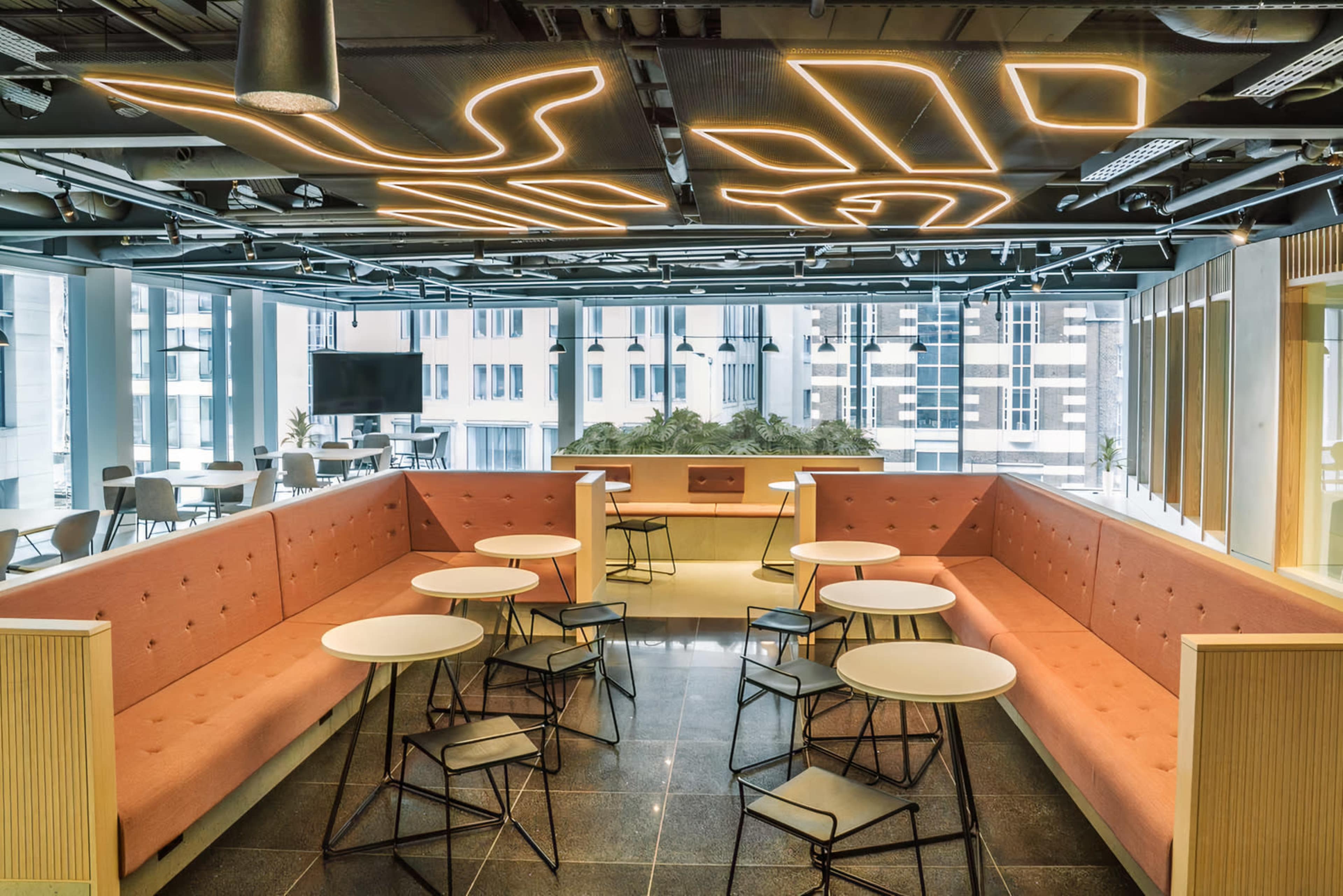 The image shows a modern lounge area with a large orange seating arrangement and round tables, illuminated by unique ceiling lights.