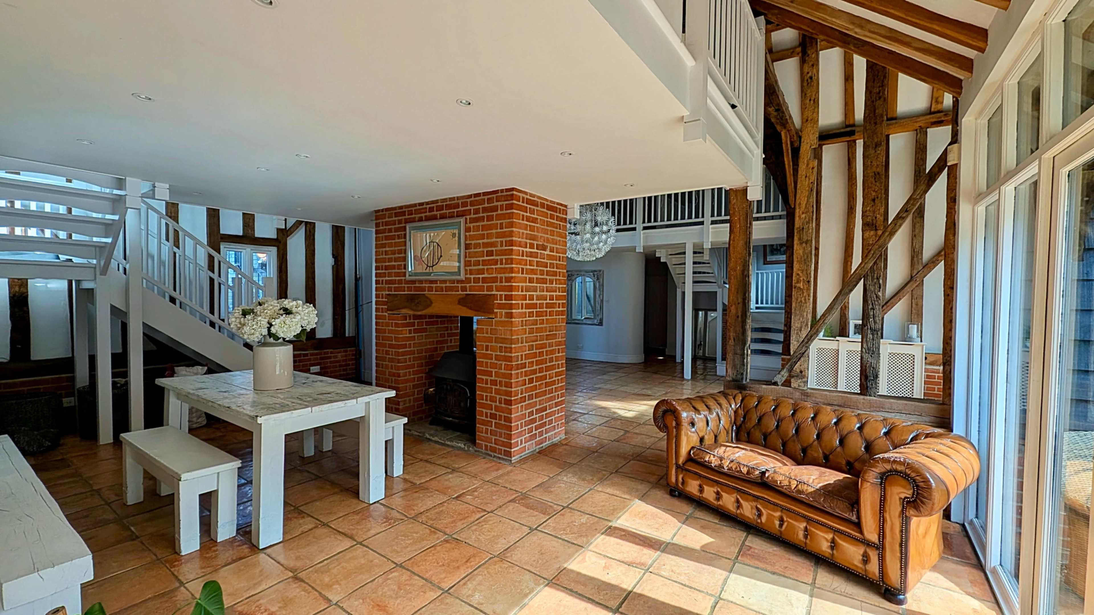 The image depicts a spacious, well-lit living area featuring a brick fireplace, a wooden dining table, a leather sofa, and exposed wooden beams.