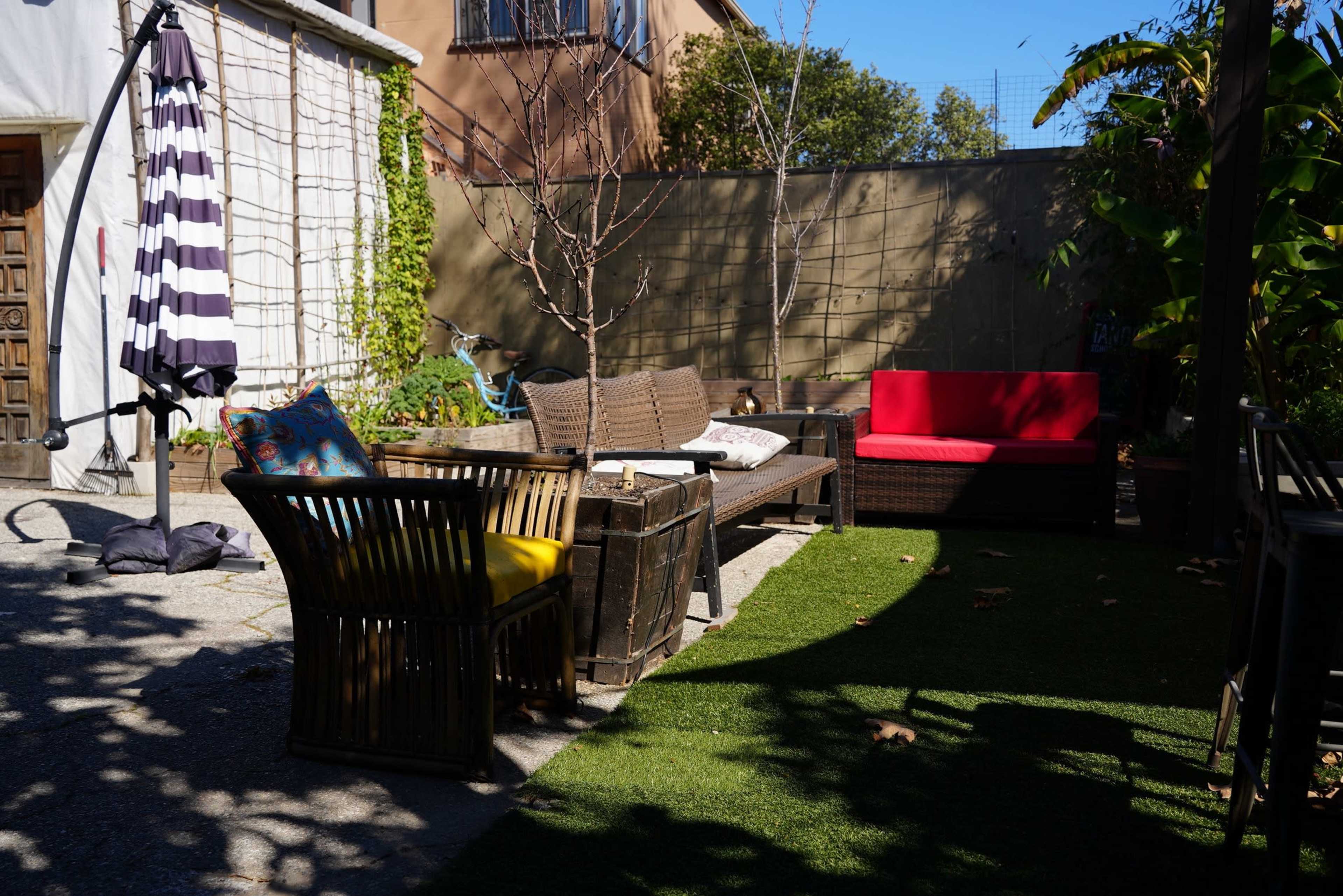 A backyard patio area with wicker furniture, including a yellow-cushioned chair, a red couch, and a table, surrounded by greenery and a twilit wall.