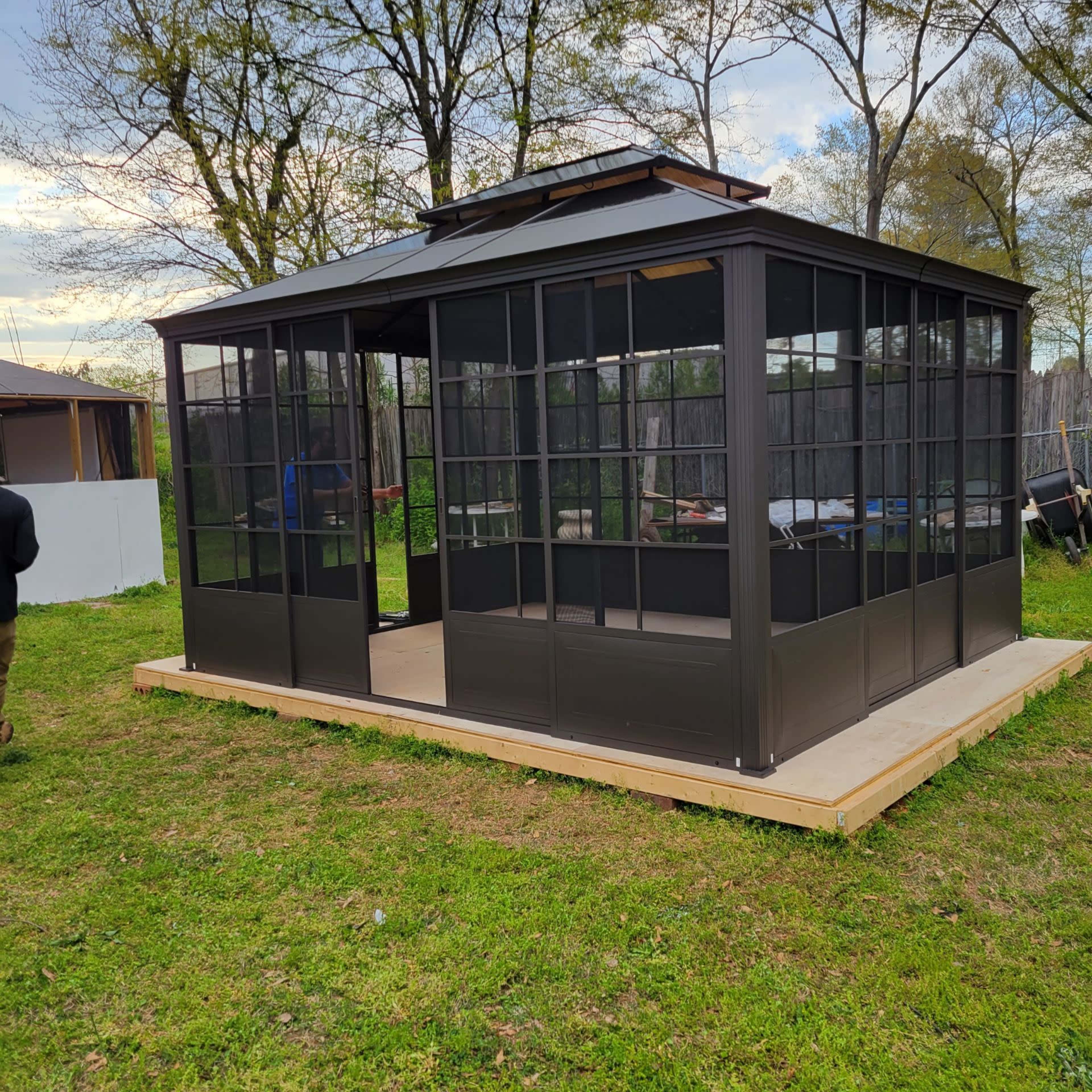 A black screened gazebo with large windows is situated on a wooden platform in a grassy area.