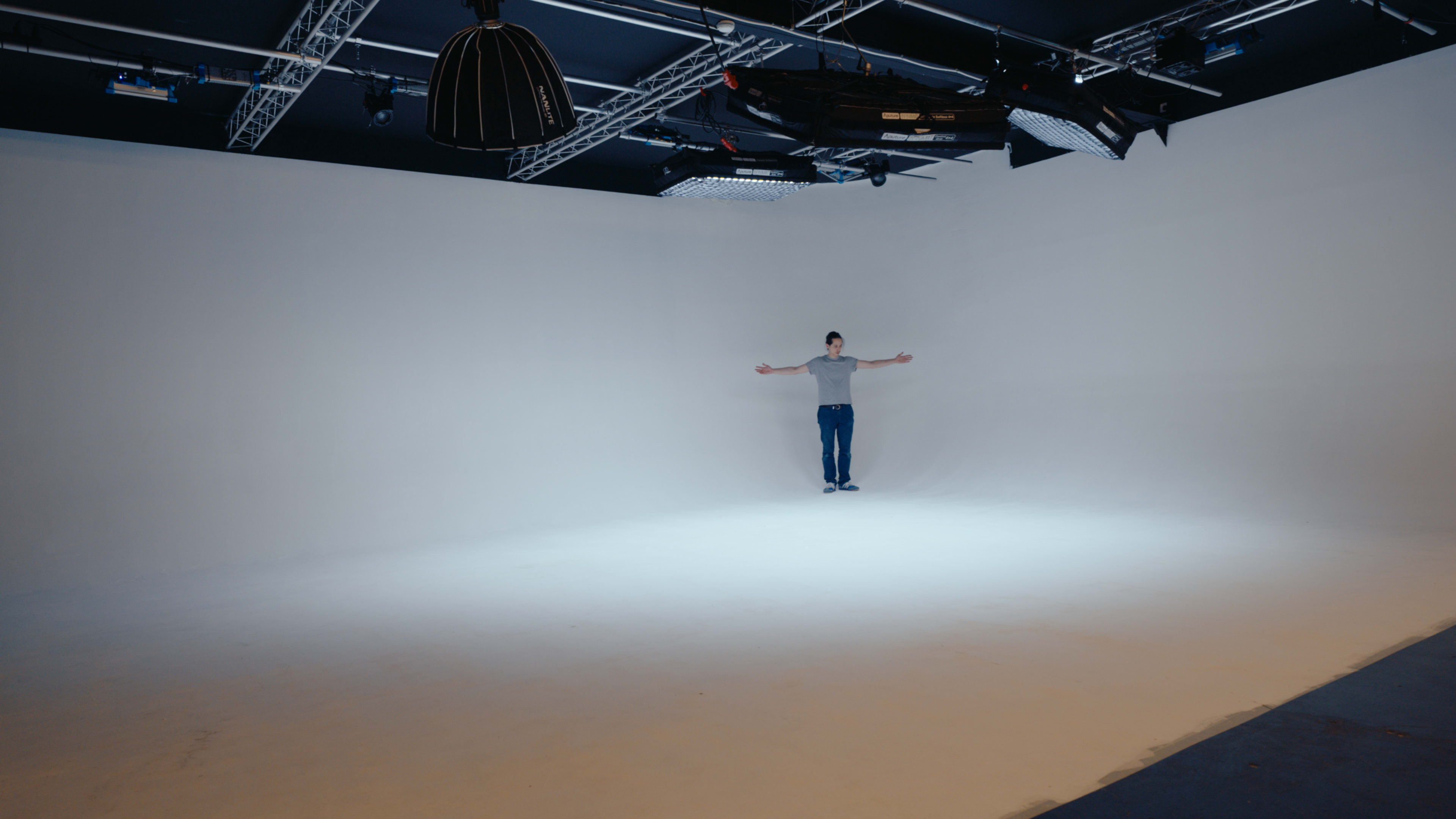 A person stands with arms outstretched in a spacious, brightly lit studio with a plain white backdrop.