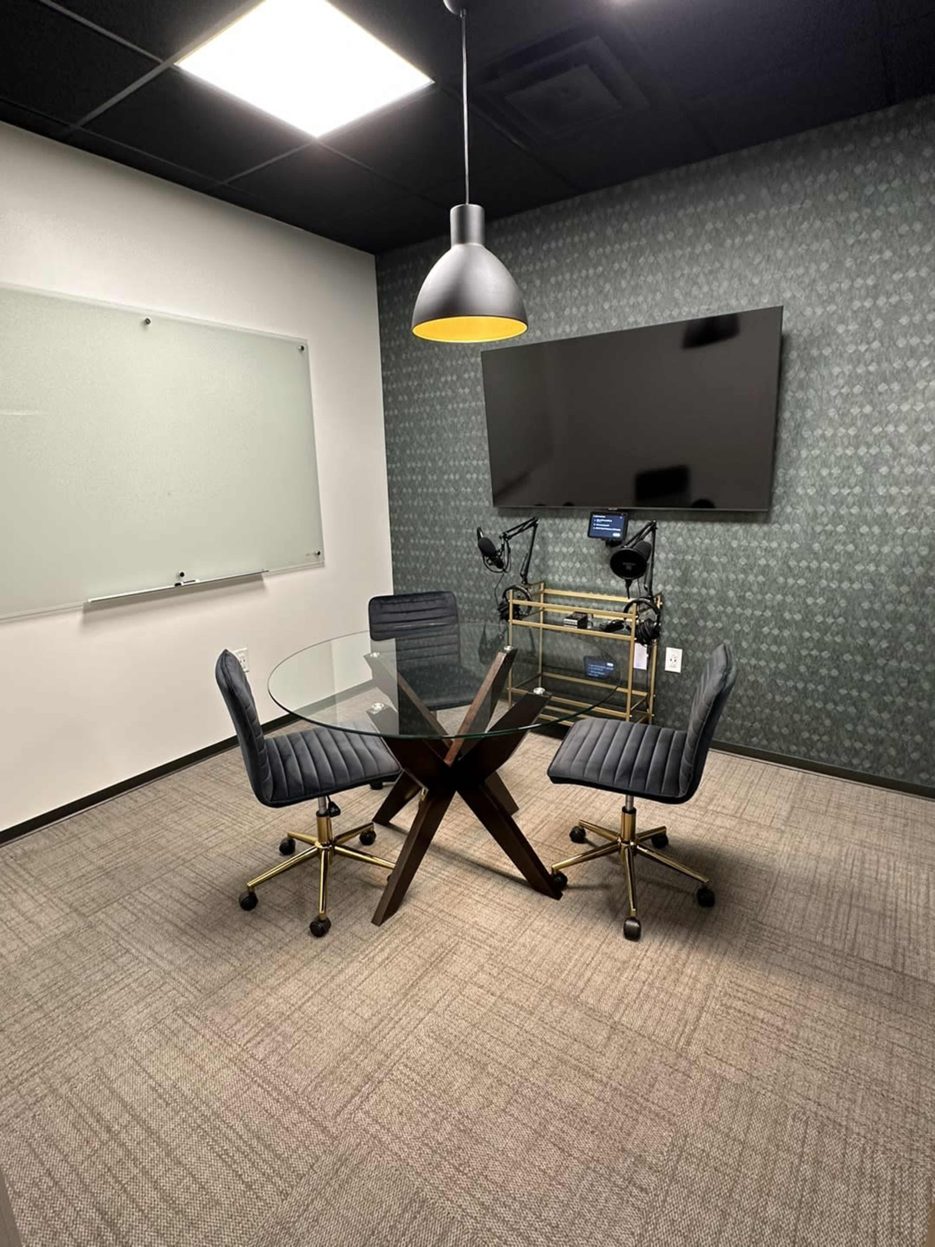 A small conference room featuring a glass table with four chairs, a wall-mounted television, and a whiteboard.