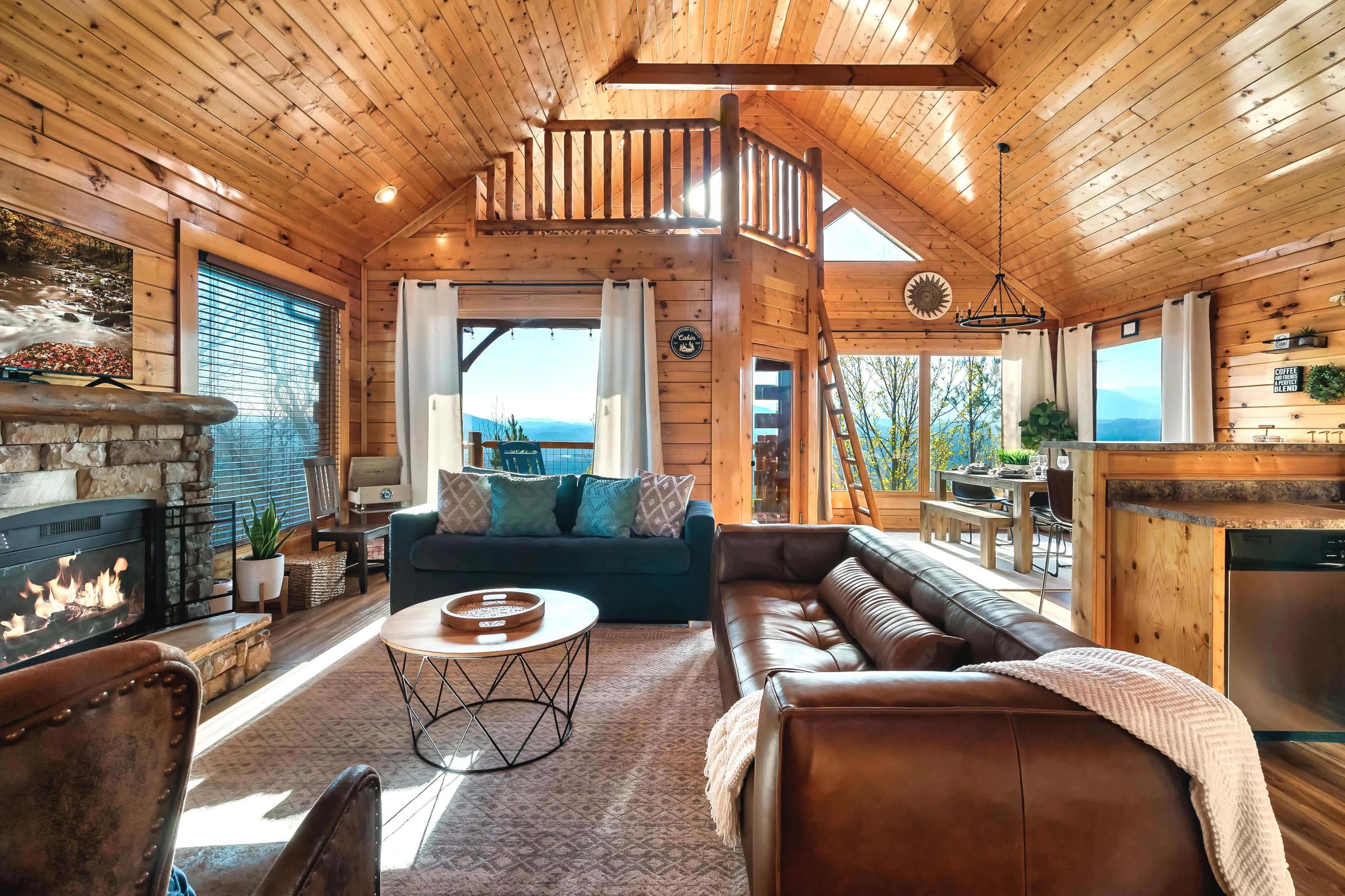 The image shows a cozy wooden cabin interior with a stone fireplace, large windows, a green sofa, and a dining area.