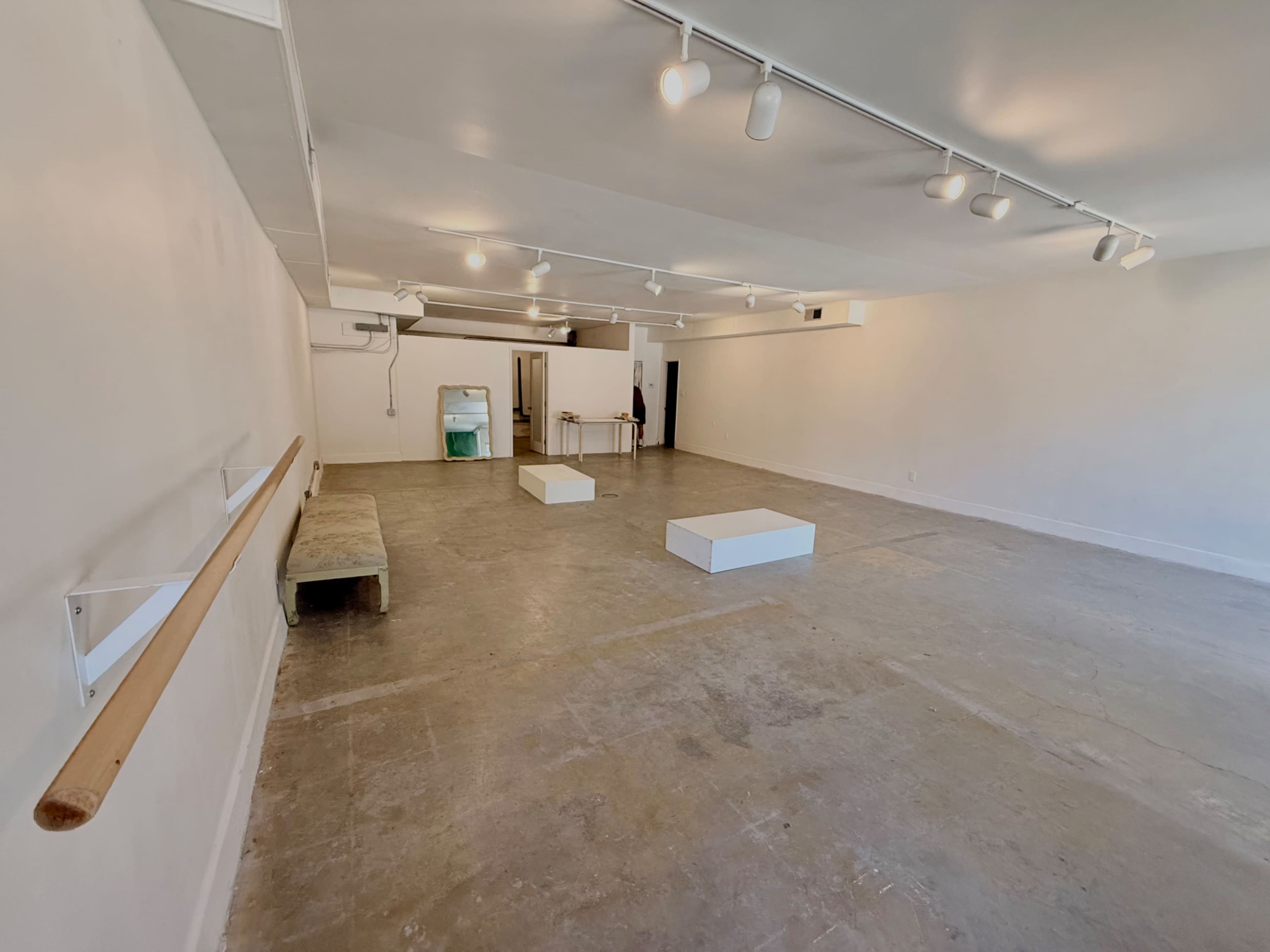 The image shows a large, empty room with wooden flooring, two white blocks positioned in the center, a barre along one wall, and a small table near the back.