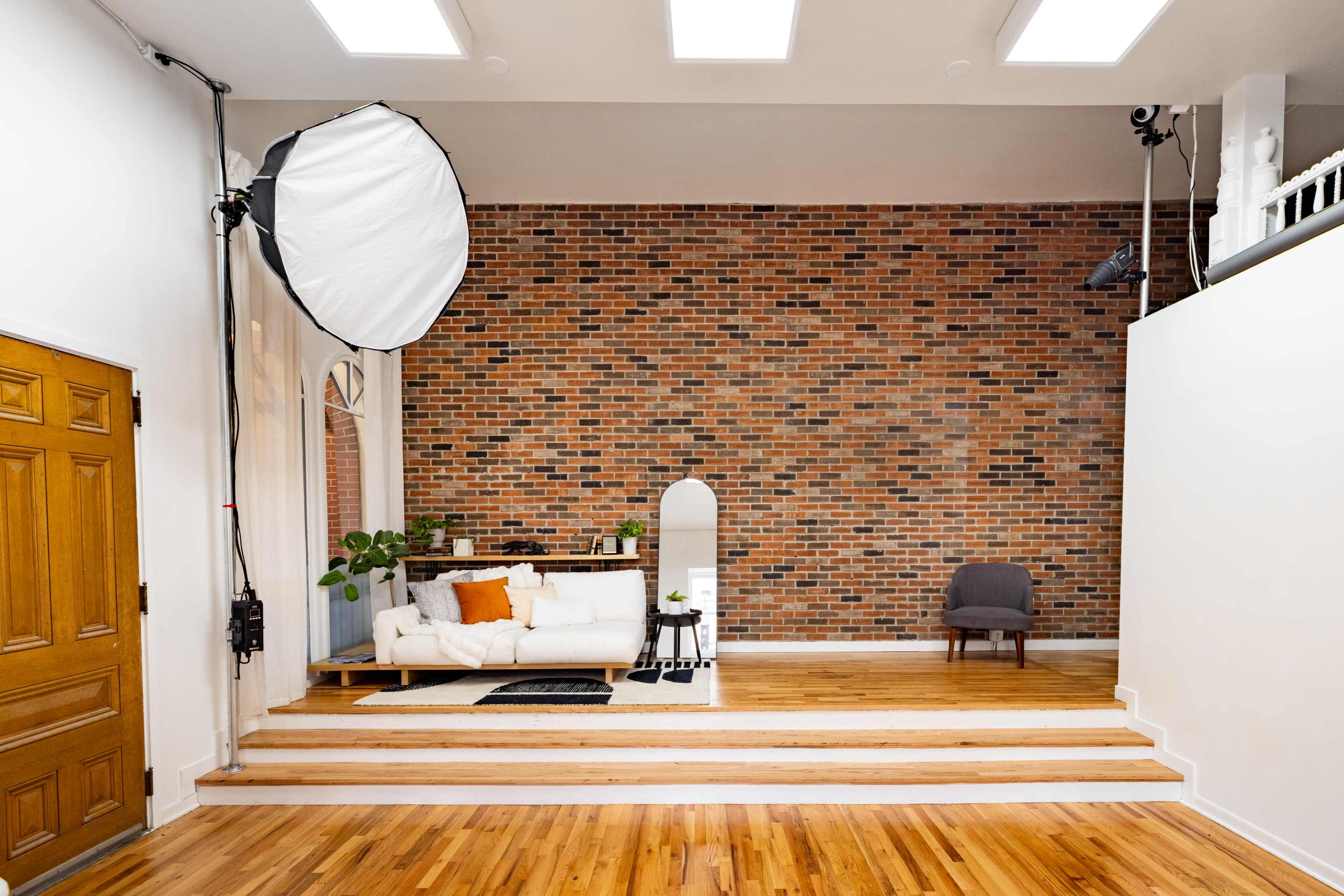 The image shows a spacious interior with a brick wall, a white couch with orange and black pillows, a round mirror, and a chair, all set on wooden flooring with a few decorative plants.