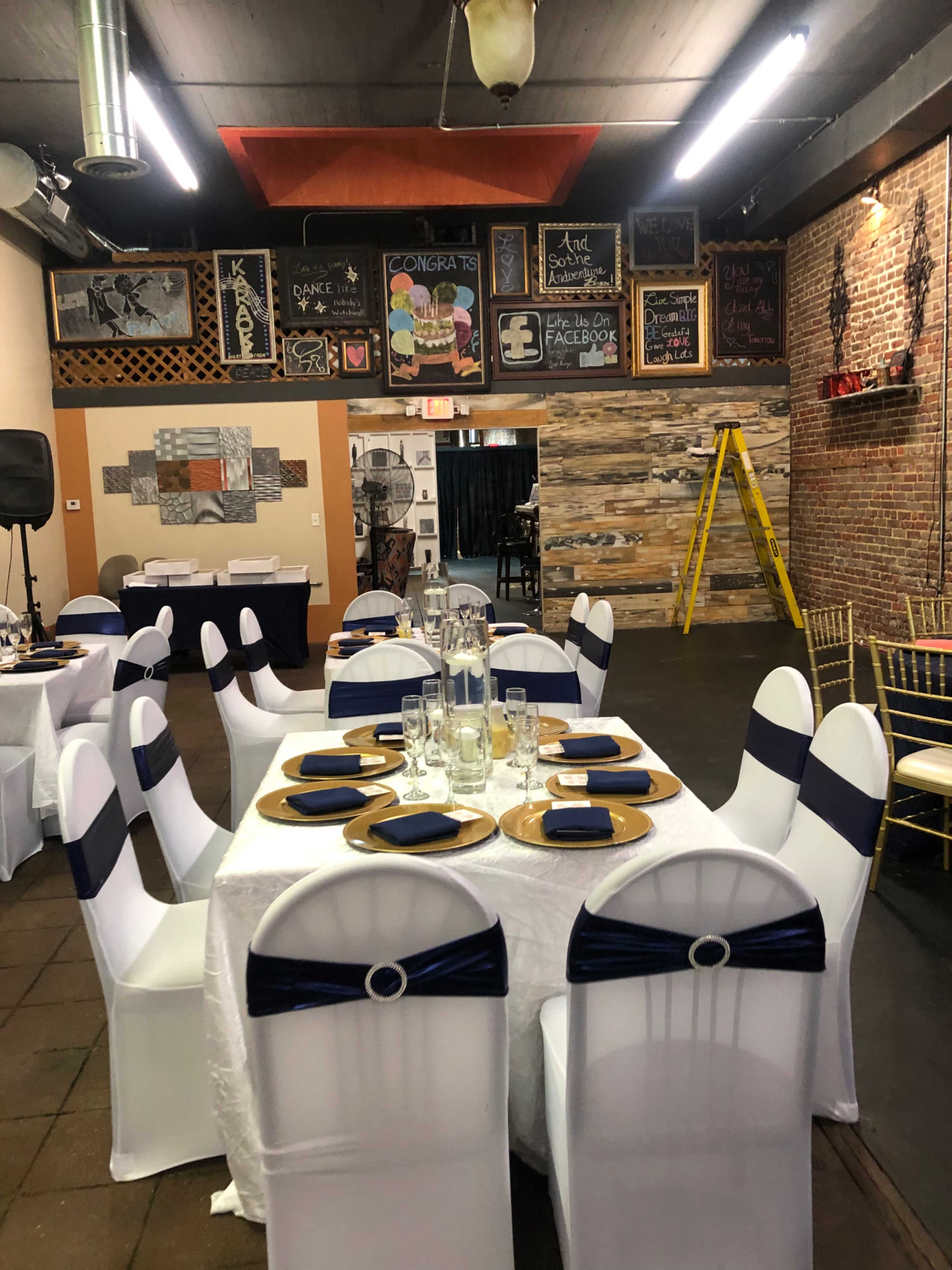 A decorated event space featuring tables set with gold chargers and dark blue napkins, surrounded by exposed brick walls and various chalkboard decorations.
