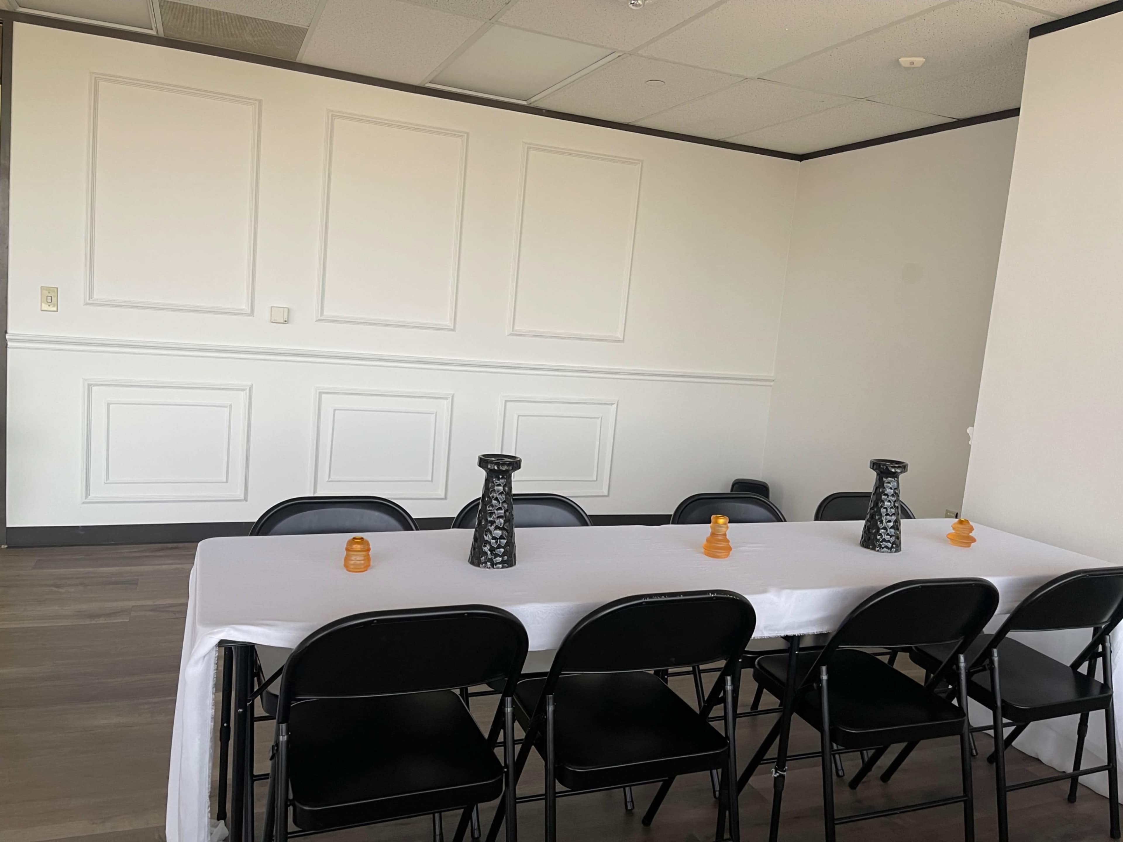 A rectangular table is set in a room with plain white walls and black chairs, featuring two vases and two small orange decorations on the table.
