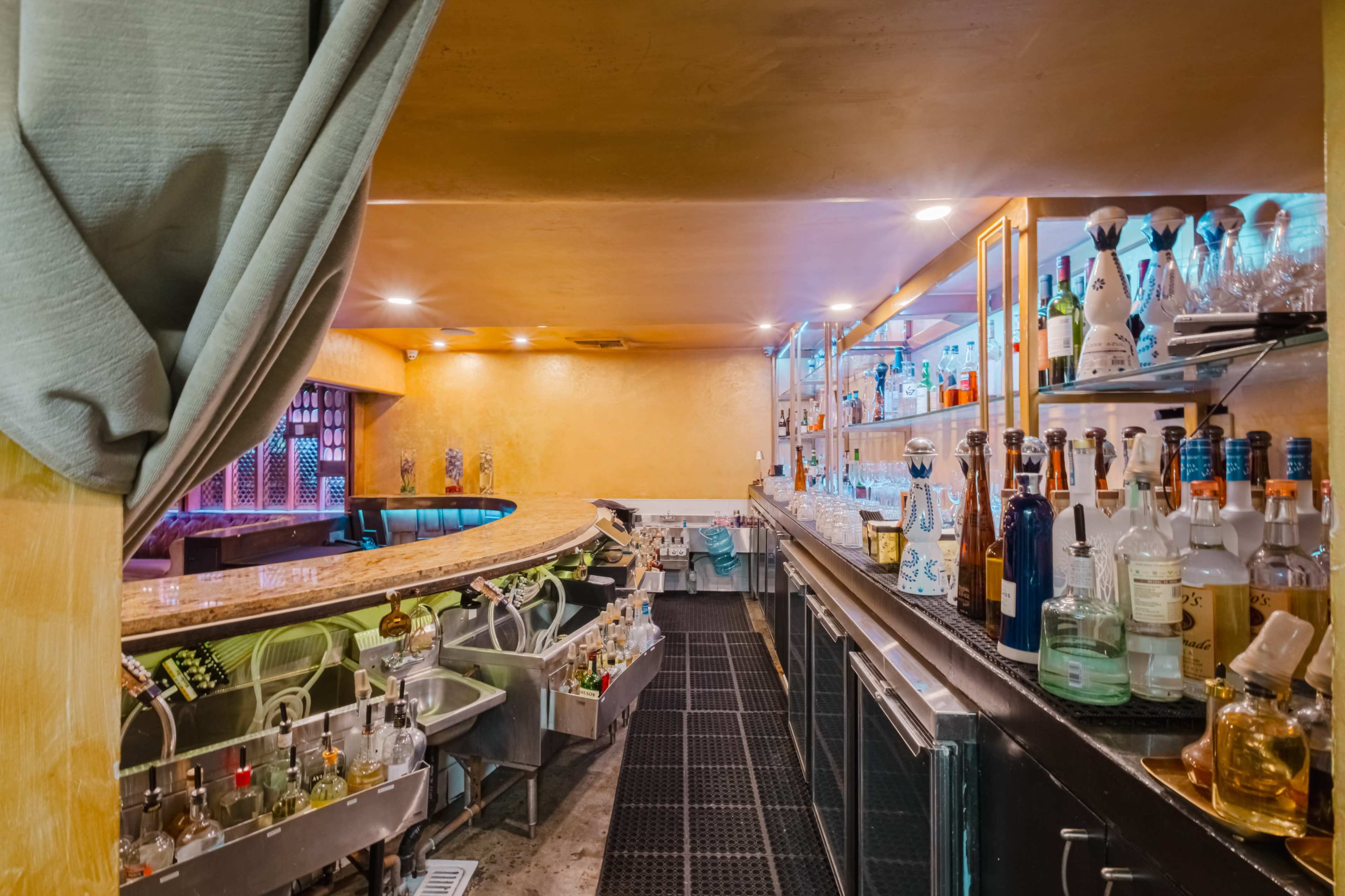 The image shows a bar area with an elongated countertop, shelves stocked with various bottles, and a well-organized display of glassware.
