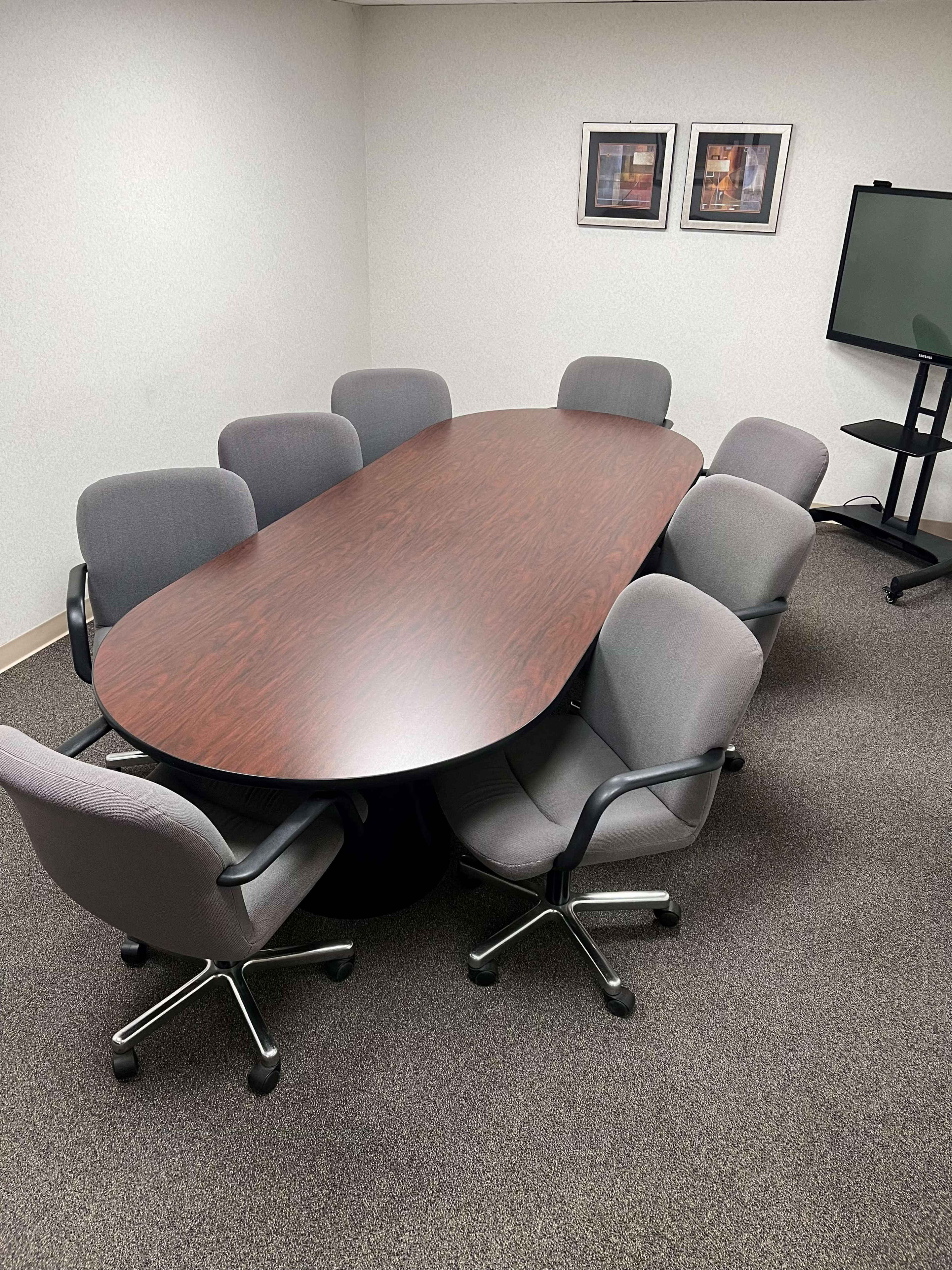 A large oval wooden conference table surrounded by eight gray office chairs is set up in a meeting room with a TV stand and framed pictures on the wall.